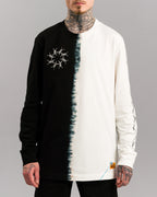 Frontalansicht eines Longsleeves mit schwarzer linker und naturweißer rechter Seite. In der Mitte verläuft eine unregelmäßige, tie-dye-artige Nahtlinie. Linke Brustseite zeigt ein kreisförmiges Stacheldraht-Stickmotiv. Am unteren rechten Saum ist ein orangefarbenes Label mit blauer Ziernaht angebracht.