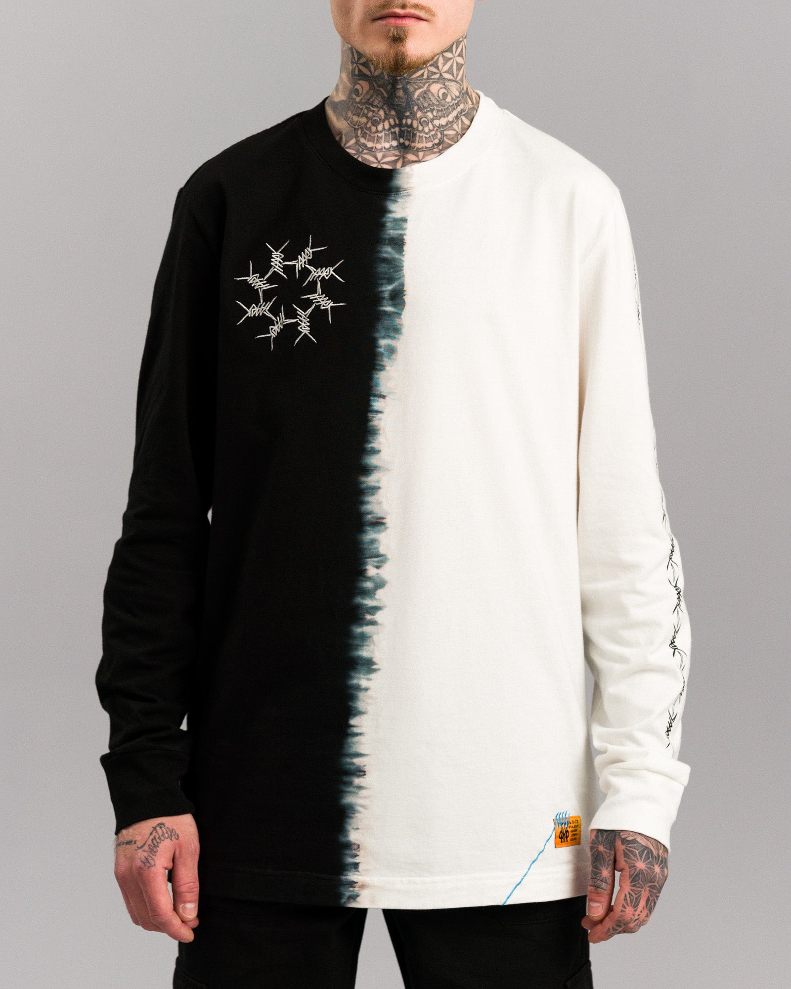 Frontalansicht eines Longsleeves mit schwarzer linker und naturweißer rechter Seite. In der Mitte verläuft eine unregelmäßige, tie-dye-artige Nahtlinie. Linke Brustseite zeigt ein kreisförmiges Stacheldraht-Stickmotiv. Am unteren rechten Saum ist ein orangefarbenes Label mit blauer Ziernaht angebracht.