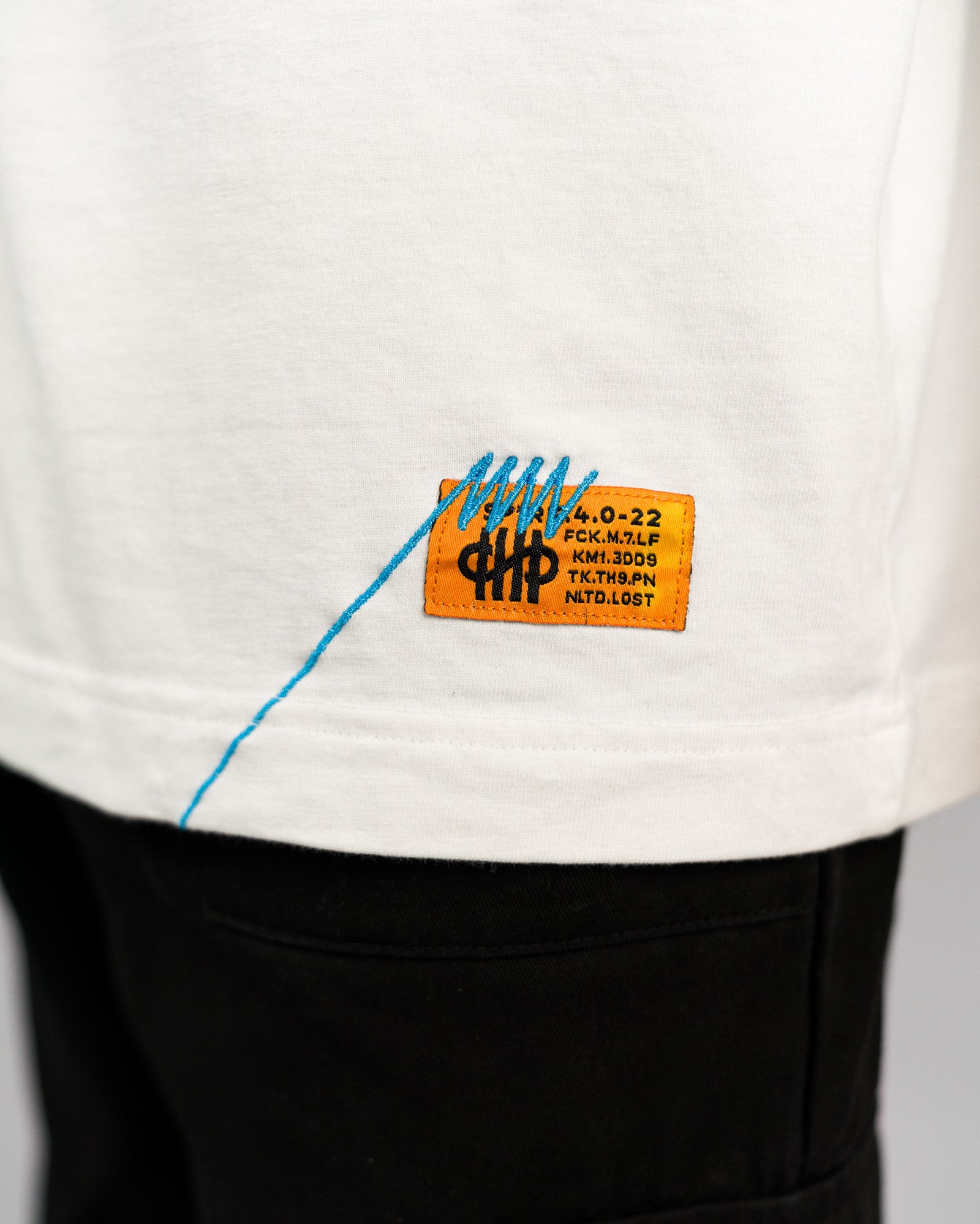 Detailaufnahme des Saumlabels: orangefarbenes Patch mit schwarzer Aufschrift und blauer Sticklinie, die scheinbar quer über das Patch „genäht“ wurde.