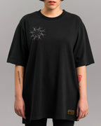 Locker geschnittenes, schwarzes T-Shirt mit feiner Stickerei auf der linken Brustseite. Die Stickerei zeigt ein kreisförmiges Muster aus grauen Stacheldrahtmotiven. Das Shirt endet locker über der Hüfte.
