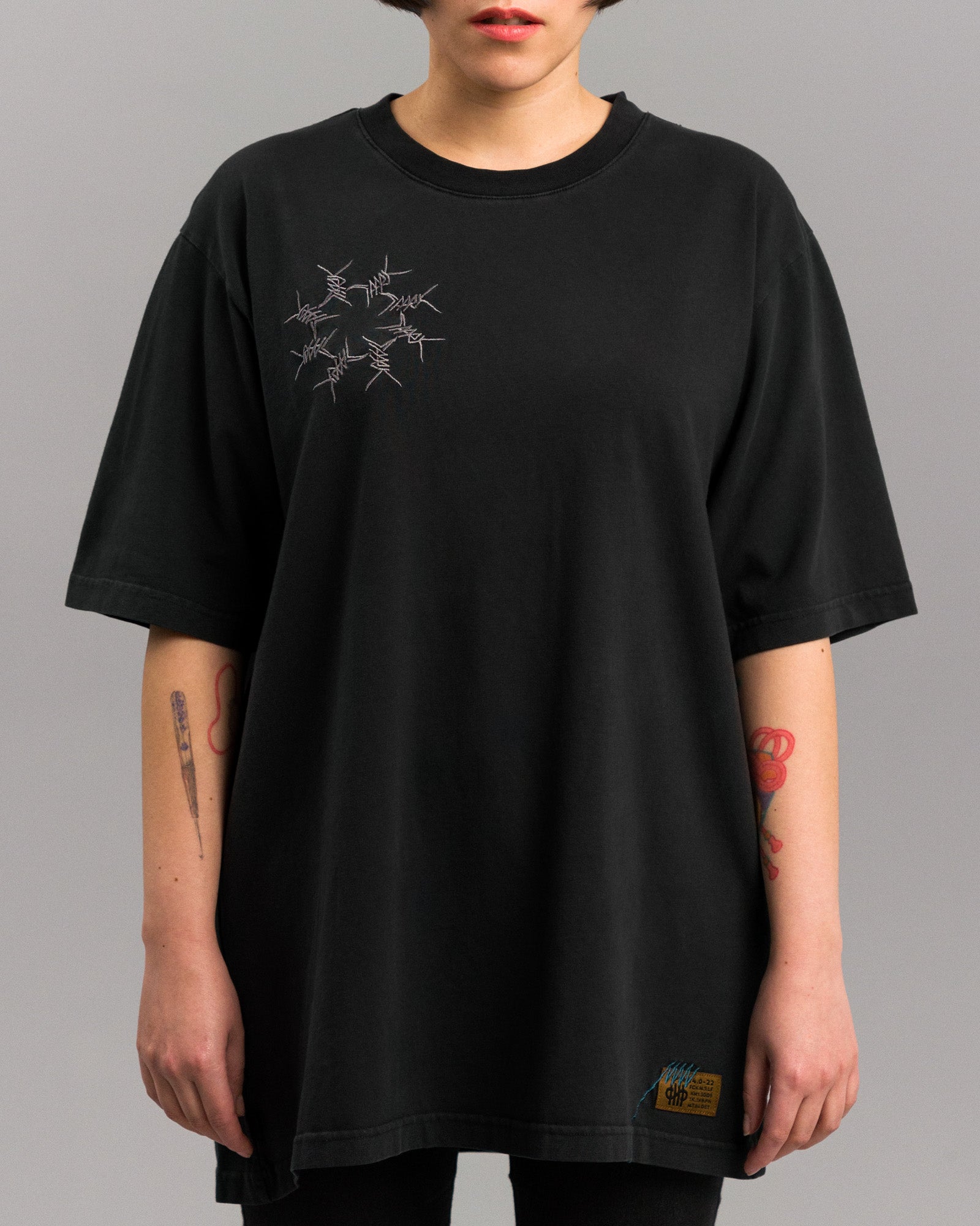 Locker geschnittenes, schwarzes T-Shirt mit feiner Stickerei auf der linken Brustseite. Die Stickerei zeigt ein kreisförmiges Muster aus grauen Stacheldrahtmotiven. Das Shirt endet locker über der Hüfte.