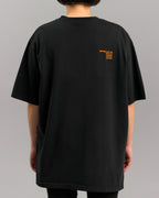Rückseite eines schwarzen, locker sitzenden T-Shirts mit kleinem orangen Textprint im rechten Schulterbereich. Der Print enthält codierte Begriffe und Zahlen. Die Ärmel sind weit geschnitten.