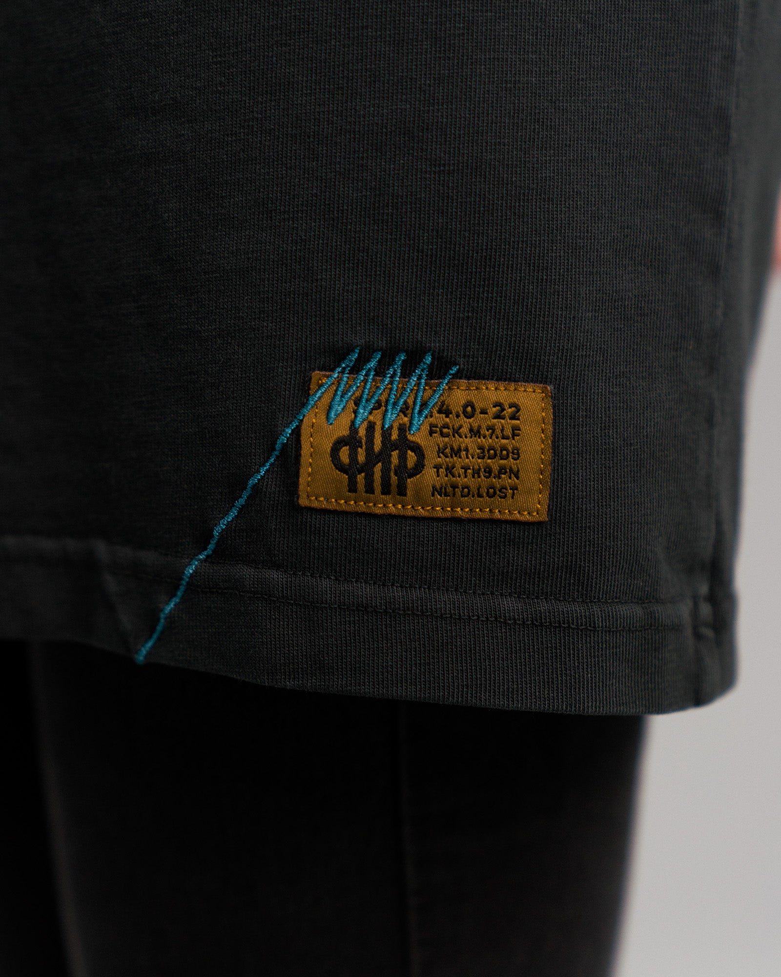 Detail des rechteckigen Markenlabels am unteren T-Shirt-Saum: ockerfarbener Aufnäher mit schwarzem Text in mehreren Zeilen und blauer Zierstickerei auf schwarzem Baumwollstoff.