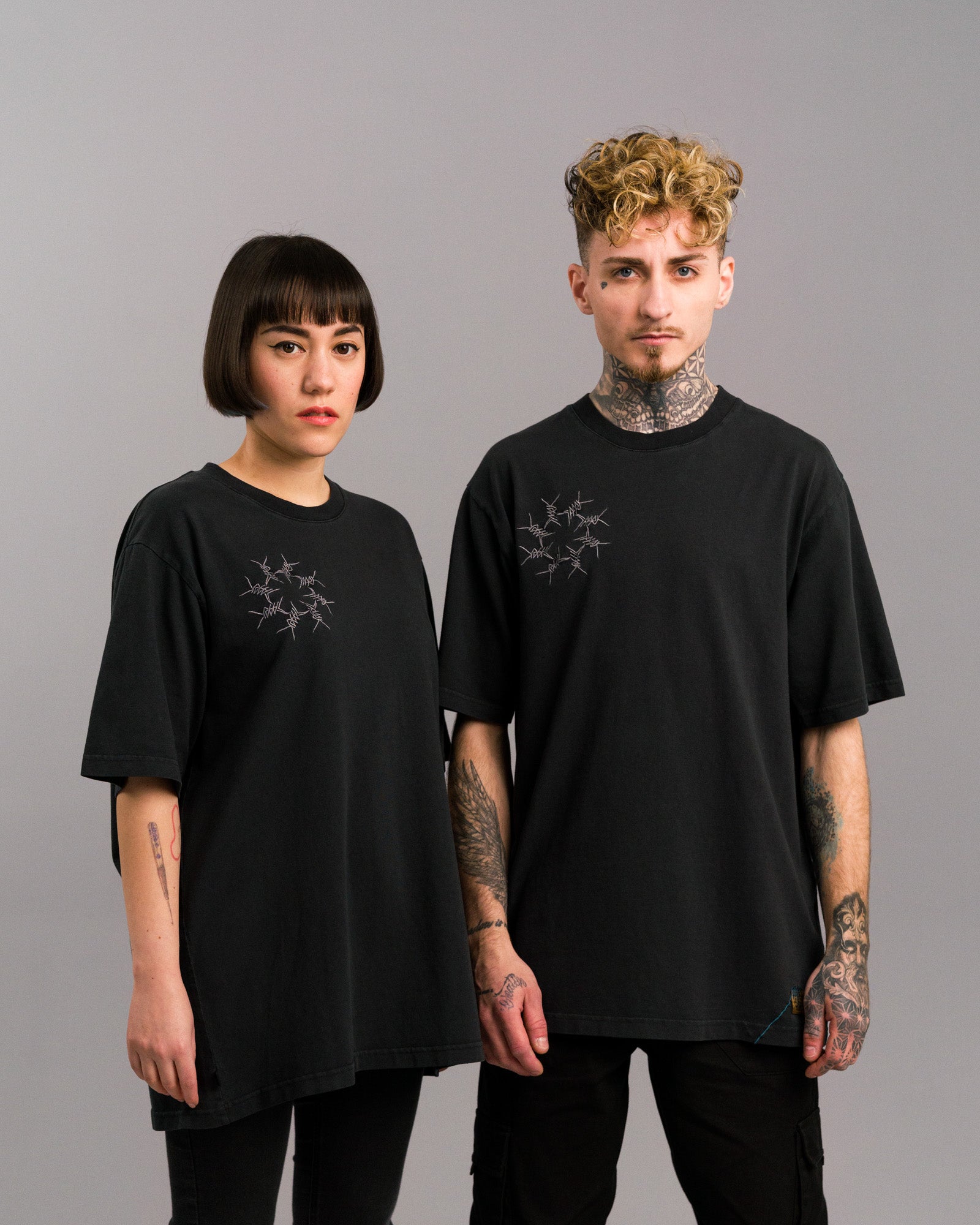 Zwei Personen tragen identische schwarze T-Shirts mit grauer Stacheldraht-Stickerei auf der linken Brust. Die Shirts sind locker geschnitten und werden mit dunklen Hosen kombiniert. Neutrale Studioaufnahme.