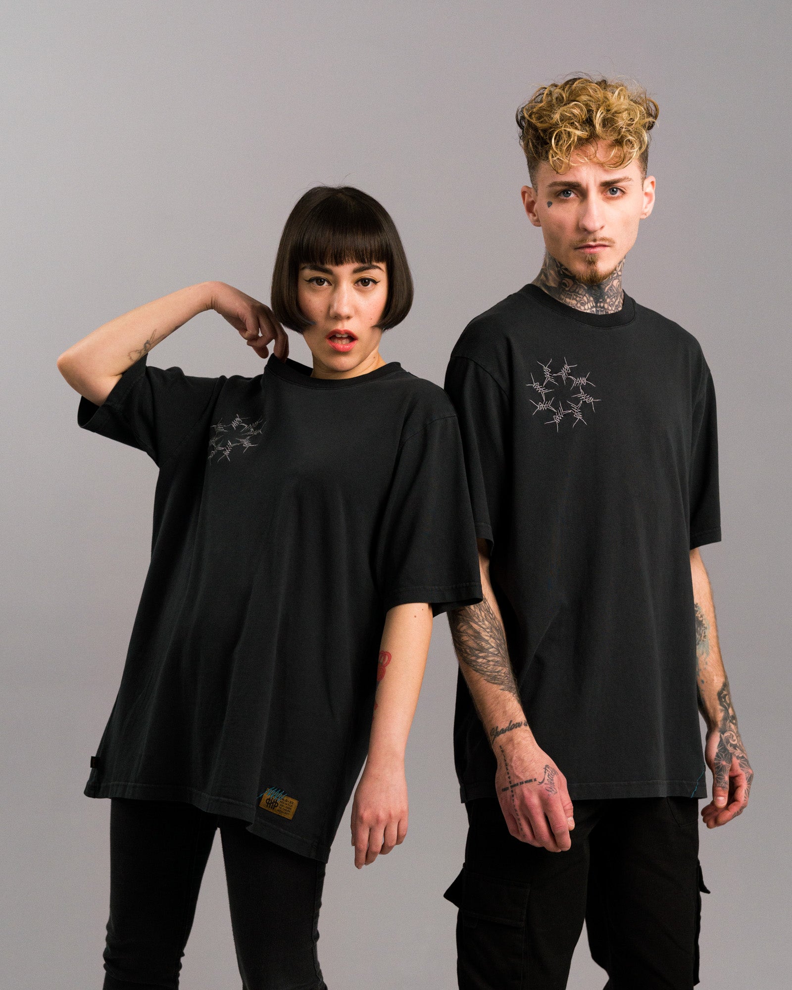 Zwei Personen tragen identische schwarze T-Shirts mit grauer Stacheldraht-Stickerei auf der linken Brust. Die Shirts sind locker geschnitten und werden mit dunklen Hosen kombiniert. Neutrale Studioaufnahme.