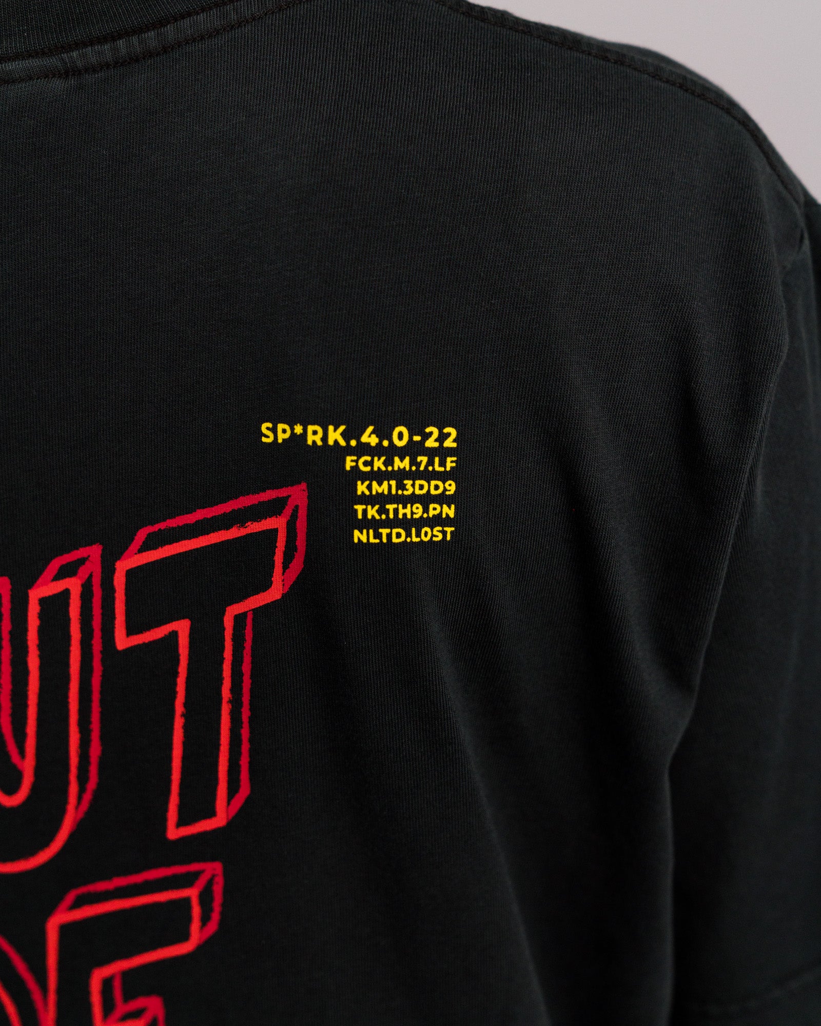 Detailaufnahme des gelben Textfelds auf dem Rücken des T-Shirts über dem „OUT OF IDEAS“-Print, mit kryptischen Codezeilen wie „SP*RK.4.0-22“ und „NLTD.LOST“.