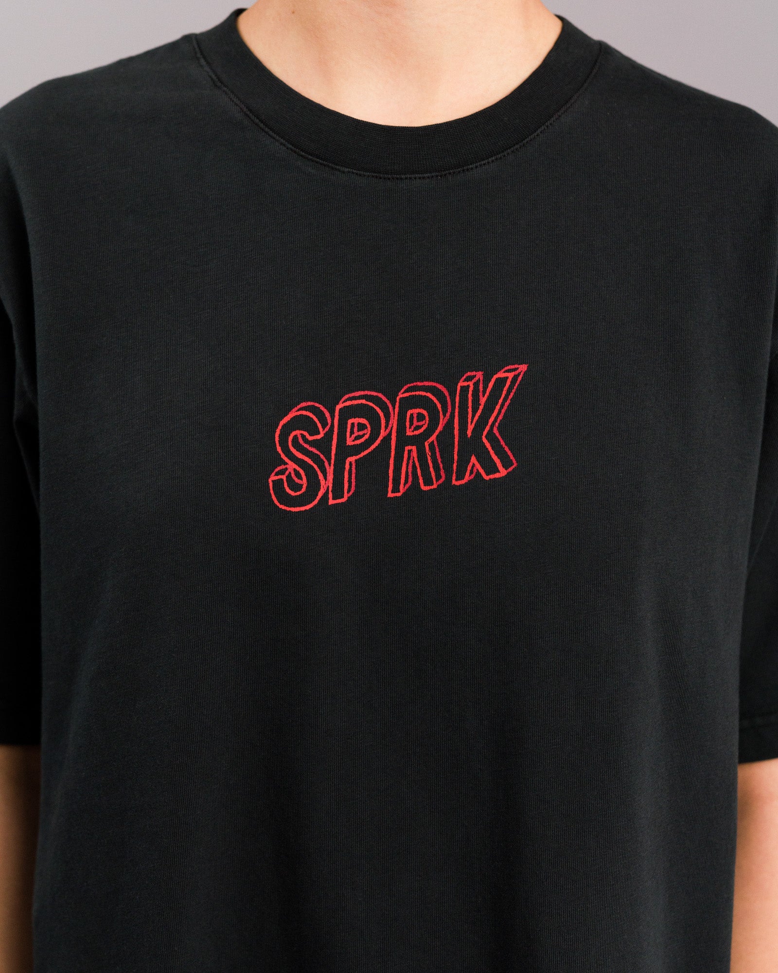 Nahaufnahme der Brustpartie eines schwarzen T-Shirts mit rotem, gesticktem „SPRK“-Schriftzug in Großbuchstaben auf der Vorderseite.