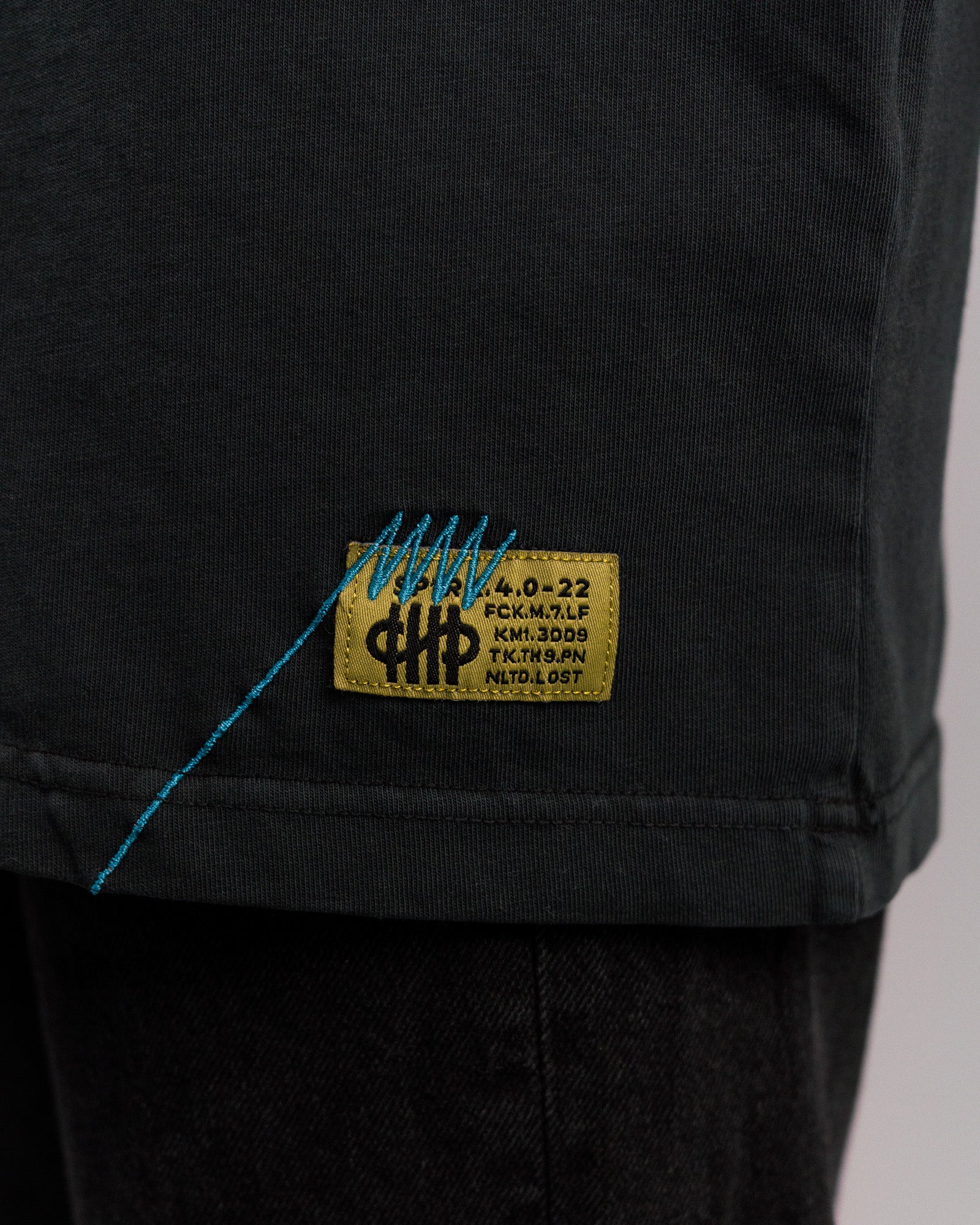 Nahaufnahme des unteren Saums des T-Shirts mit eingenähtem gelben Weblabel mit blauer Ziernaht und gelbem Textfeld „SP*RK.4.0-22“, darunter weitere Zeichenkombinationen.

