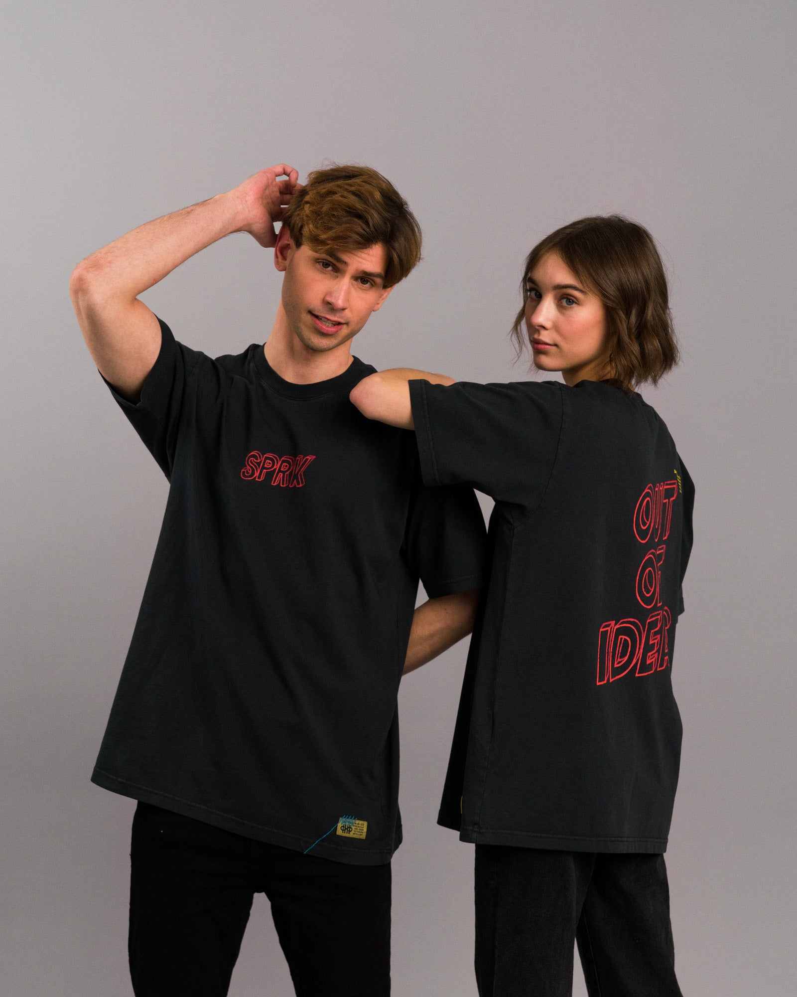 Zwei Personen nebeneinander: links mit schwarzem T-Shirt und rotem „SPRK“-Frontprint, rechts mit Rückansicht des gleichen Shirts mit „OUT OF IDEAS“-Backprint.