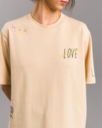 Nahaufnahme eines cremefarbenen T-Shirts mit schwarzem „LOVE“-Schriftzug auf der Brust, der durch eine gelbe Linie durchgestrichen ist. Die linke Schulter zeigt drei Smiley-Gesichter, zwei traurig, eines fröhlich. Getragen von einer Person vor grauem Hintergrund.