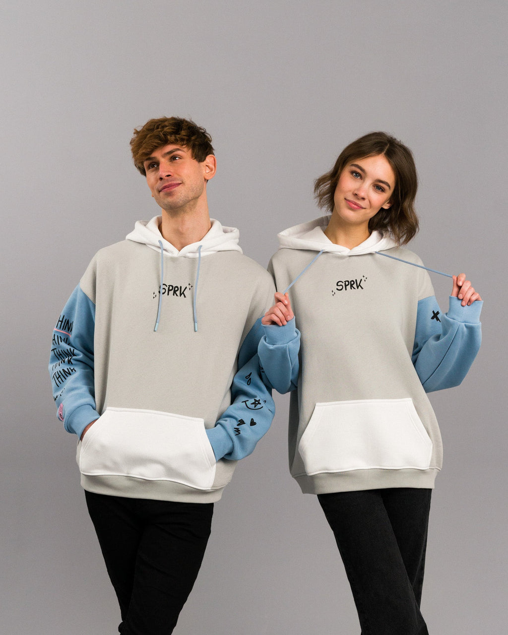 Zwei Personen stehen nebeneinander und tragen identische Hoodies mit hellem Grundton, weißen Kapuzen und hellblauen Ärmeln. Auf der Brust befindet sich ein kleiner „SPRK“-Schriftzug, auf den Ärmeln verschiedene Symbole und der Schriftzug „THINK“. Beide Personen lächeln und posieren spielerisch.