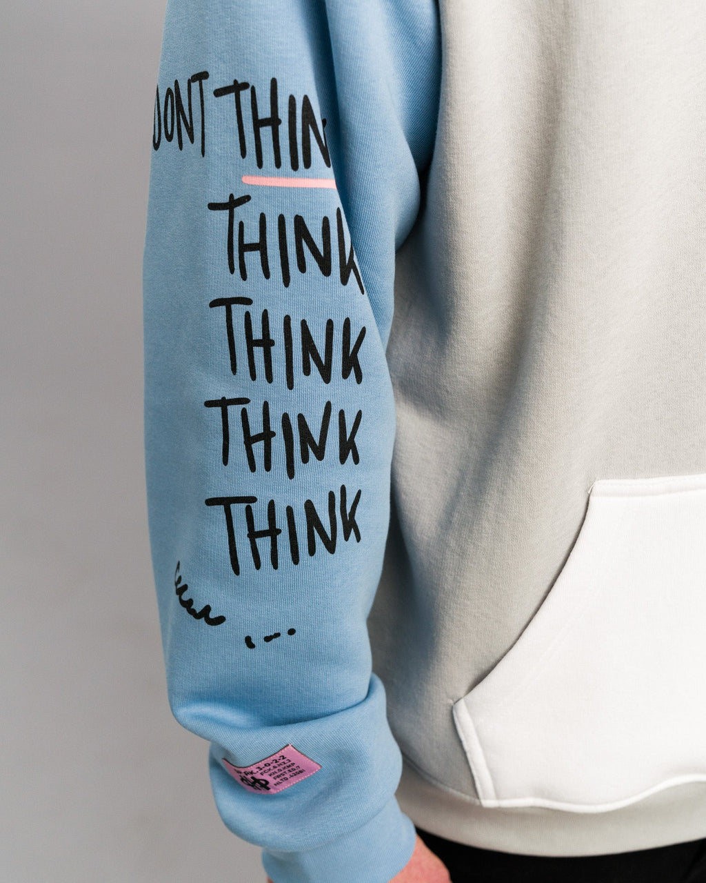 Detailansicht des rechten Ärmels. Der hellblaue Stoff zeigt den mehrfach wiederholten Schriftzug „THINK“ in schwarzer Handschriftoptik sowie ein rosafarbenes Label am Bündchen.