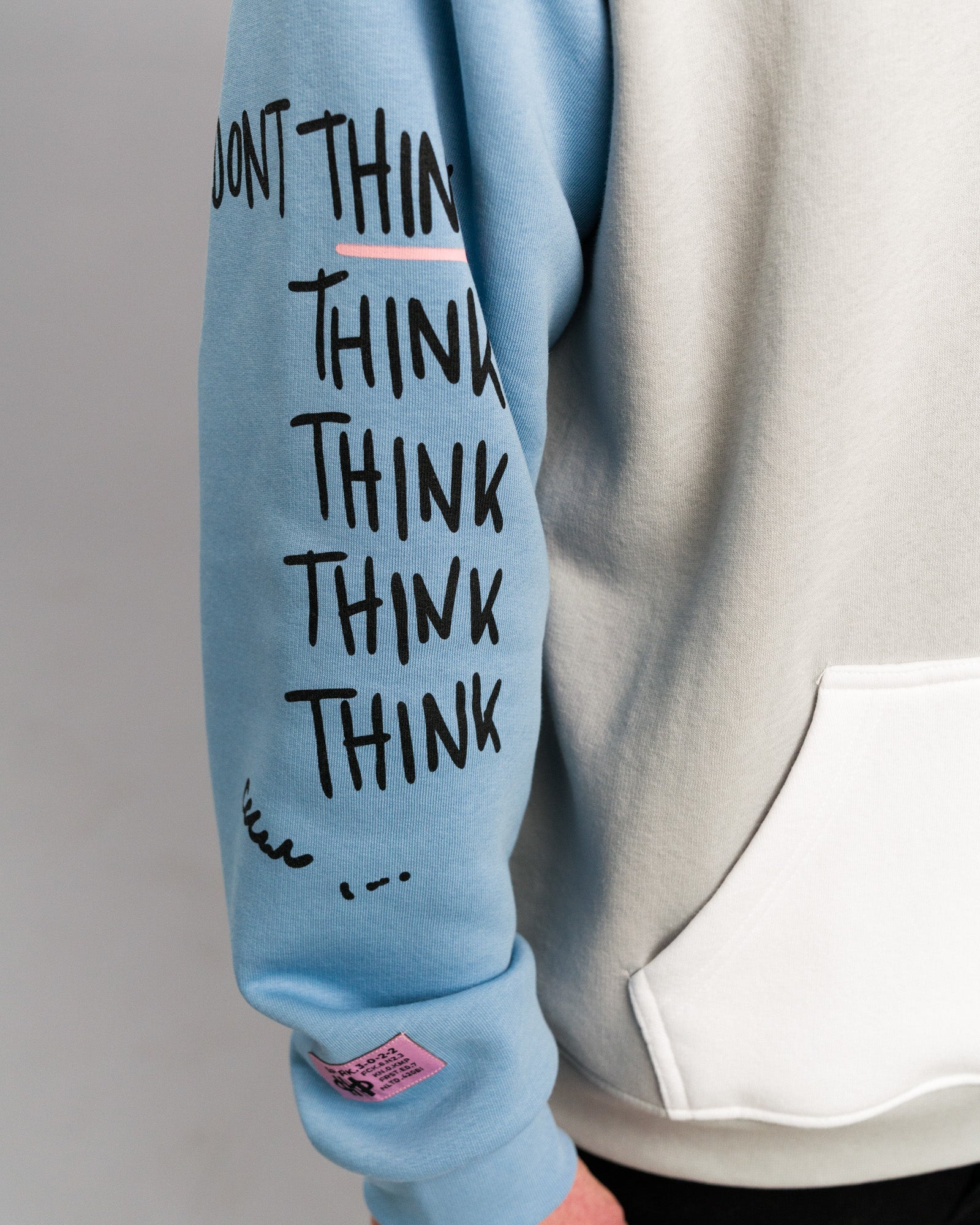 Detailansicht des rechten Ärmels. Der hellblaue Stoff zeigt den mehrfach wiederholten Schriftzug „THINK“ in schwarzer Handschriftoptik sowie ein rosafarbenes Label am Bündchen.