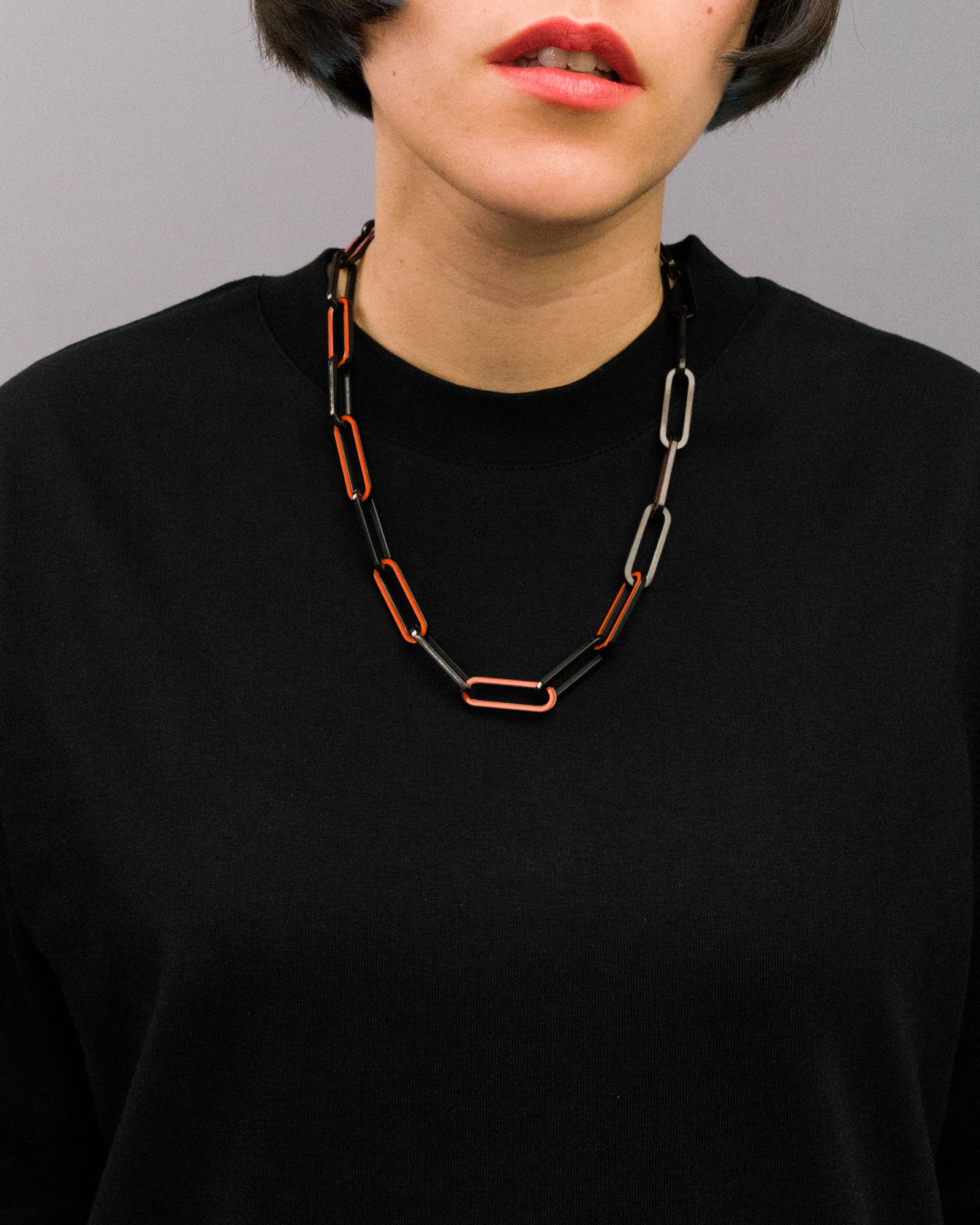 Person mit schwarzem Oberteil trägt eine markante Gliederkette mit länglichen, rechteckigen Gliedern in Schwarz und leuchtendem Orange. Die Glieder wechseln sich regelmäßig ab und bilden ein gleichmäßiges Muster um den Hals