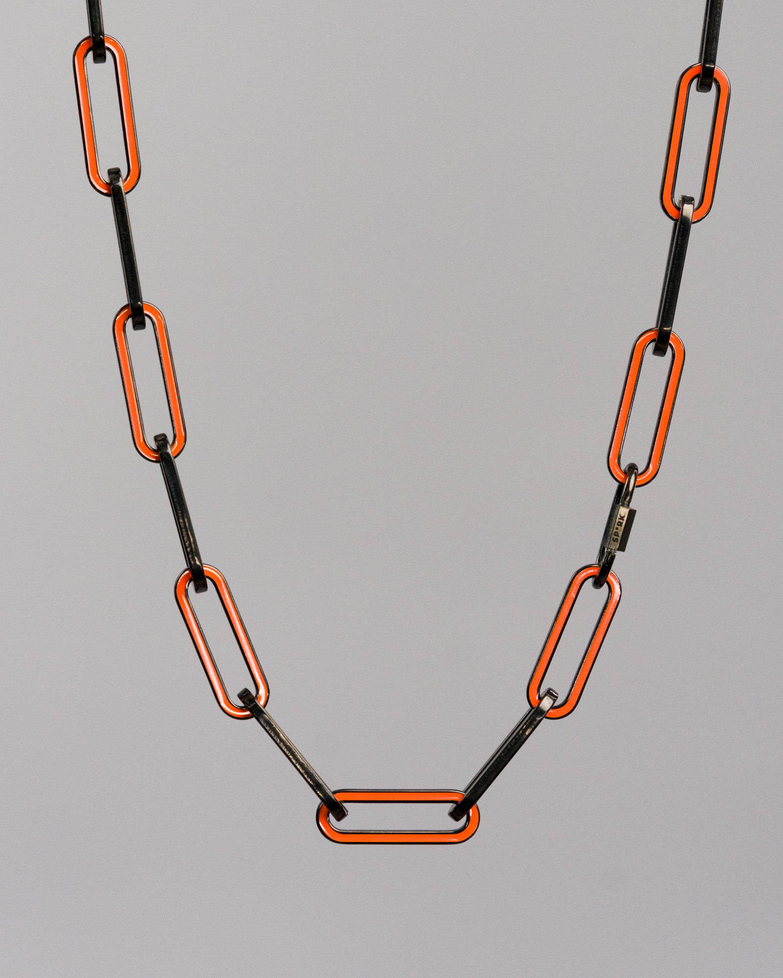 Freigestellte Nahaufnahme der Gliederkette mit länglichen, rechteckigen Elementen. Die Glieder bestehen aus schwarzem und orangenem Metall und sind abwechselnd angeordnet.

