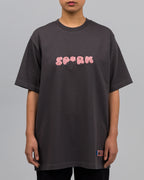 Frontalansicht einer stehenden Person in einem dunkelgrauen T-Shirt. In der Mitte der Brust ist das Wort „SP*RK“ in einer flauschig wirkenden, rosa Chenille-Optik aufgedruckt. Unten links ist ein gewebtes Label mit blauen Nähten befestigt.