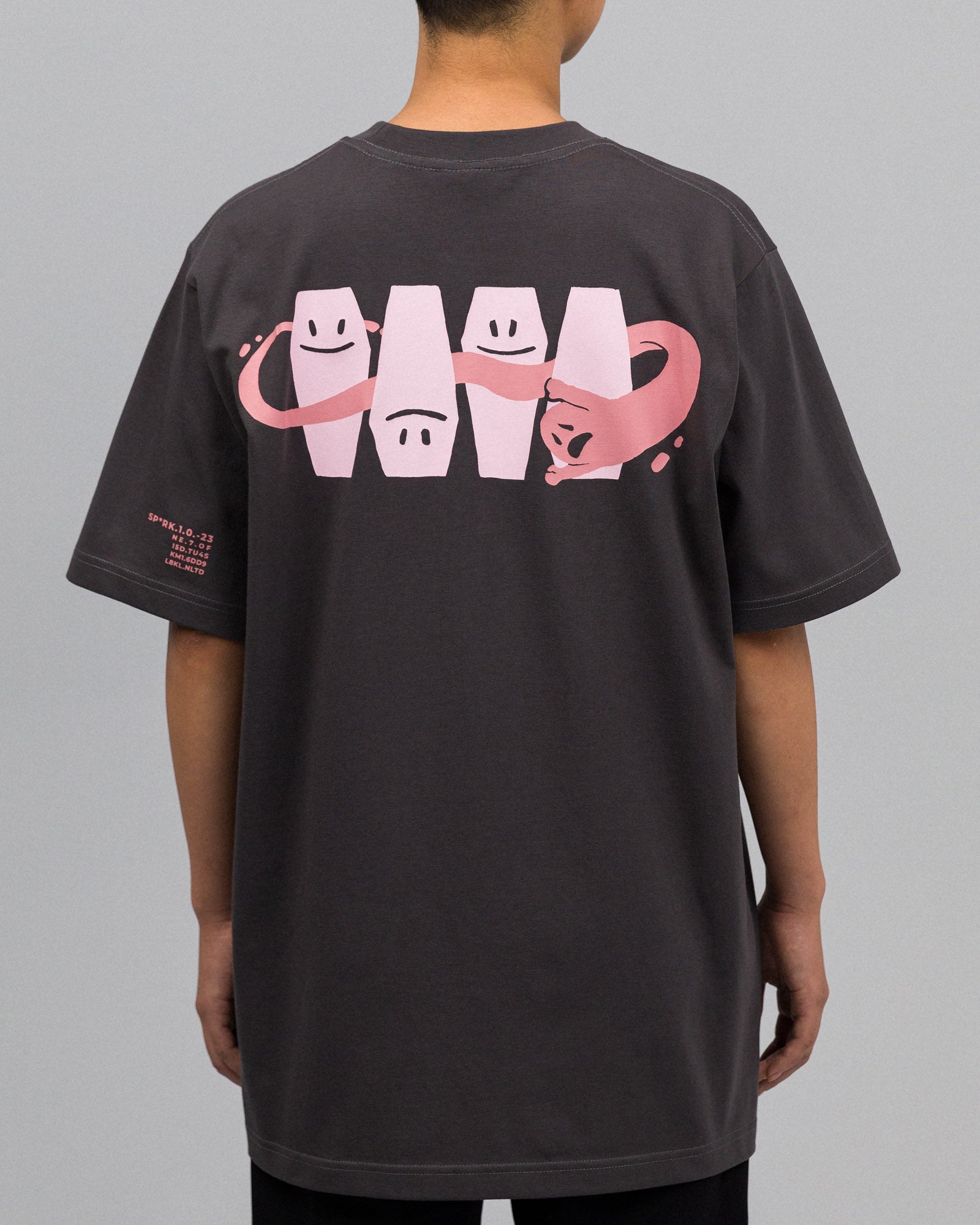 Rückansicht einer stehenden Person im dunkelgrauen T-Shirt. Zu sehen ist der großflächige Rückendruck mit den vier rosa Särgen mit Gesichtsausdrücken und dem durchlaufenden rosa Band in Form einer Katze. Auf dem linken Ärmel befindet sich zusätzlich ein kleiner rosa Schriftzug.