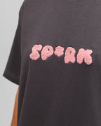 Nahaufnahme des Frontprints auf dem T-Shirt: Die Buchstaben „SP*RK“ erscheinen in plastisch-texturierter, rosa Plüschoptik auf dunkelgrauem Stoff.