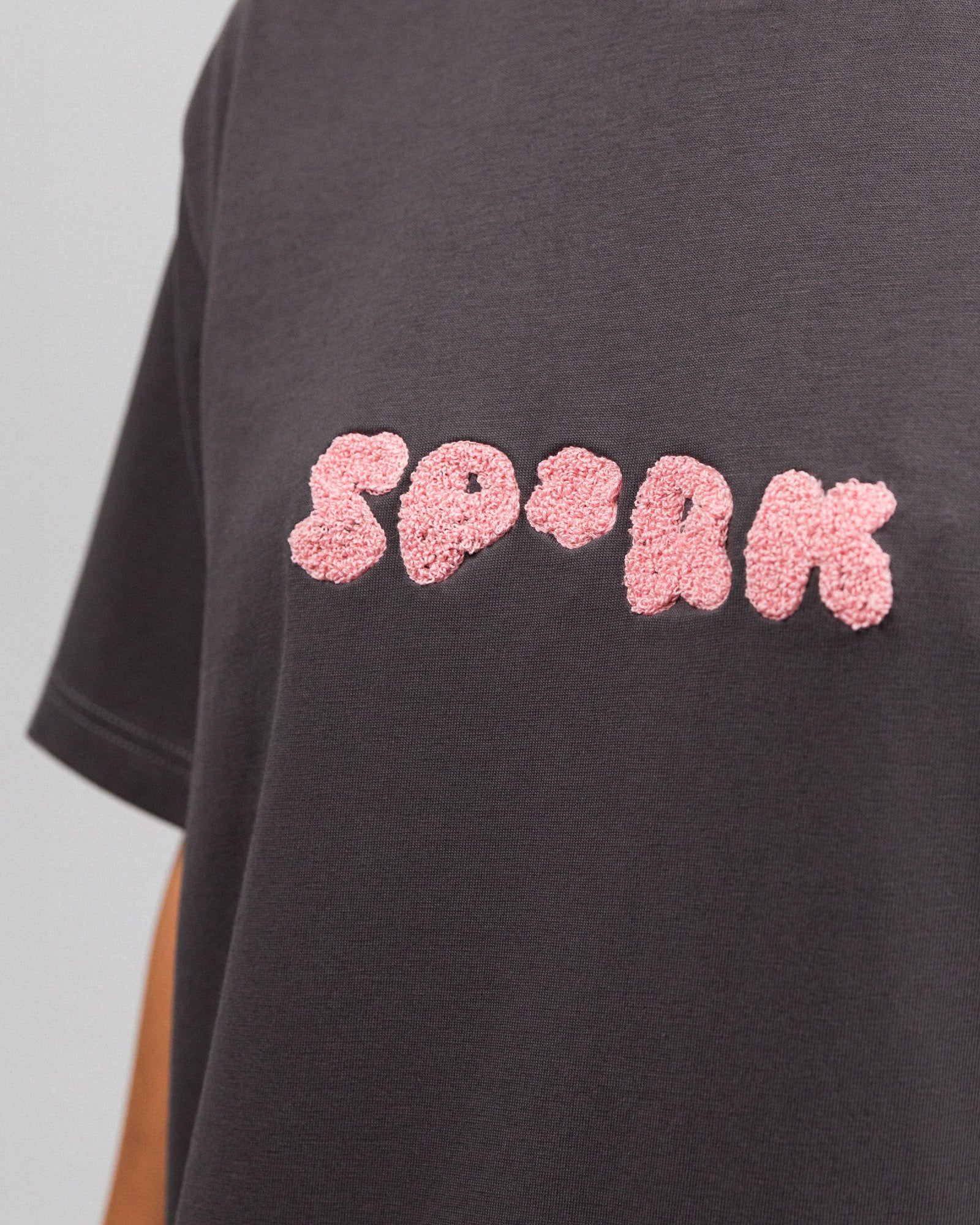 Nahaufnahme des Frontprints auf dem T-Shirt: Die Buchstaben „SP*RK“ erscheinen in plastisch-texturierter, rosa Plüschoptik auf dunkelgrauem Stoff.