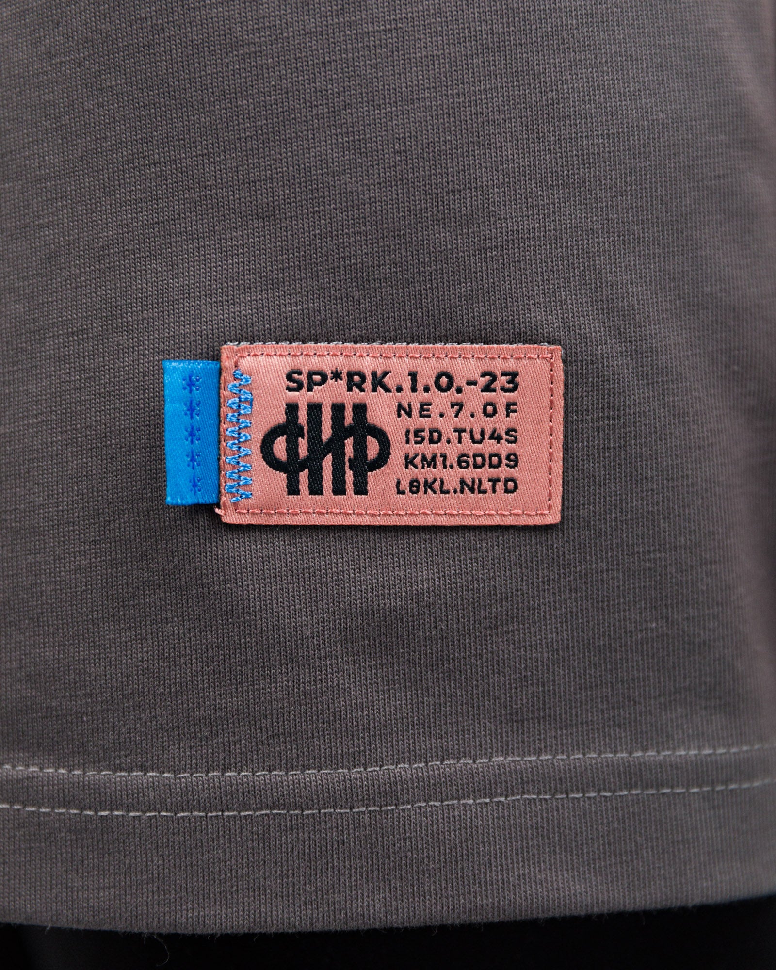 Detail des gewebten Etiketts am unteren Rand des T-Shirts. Das Label zeigt die Typografie „SP*RK.1.0.–23“ sowie weitere Zahlen-Buchstabenkombinationen auf rosa Untergrund mit blauer Naht.