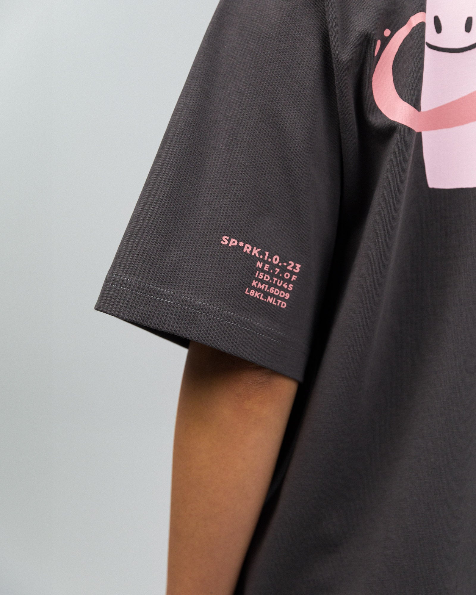 Nahaufnahme des linken T-Shirt-Ärmels mit kleinem rosa Druck: „SP*RK.1.0.–23“, gefolgt von mehreren Zahlen und Buchstabencodes, die technisch oder codiert wirken.