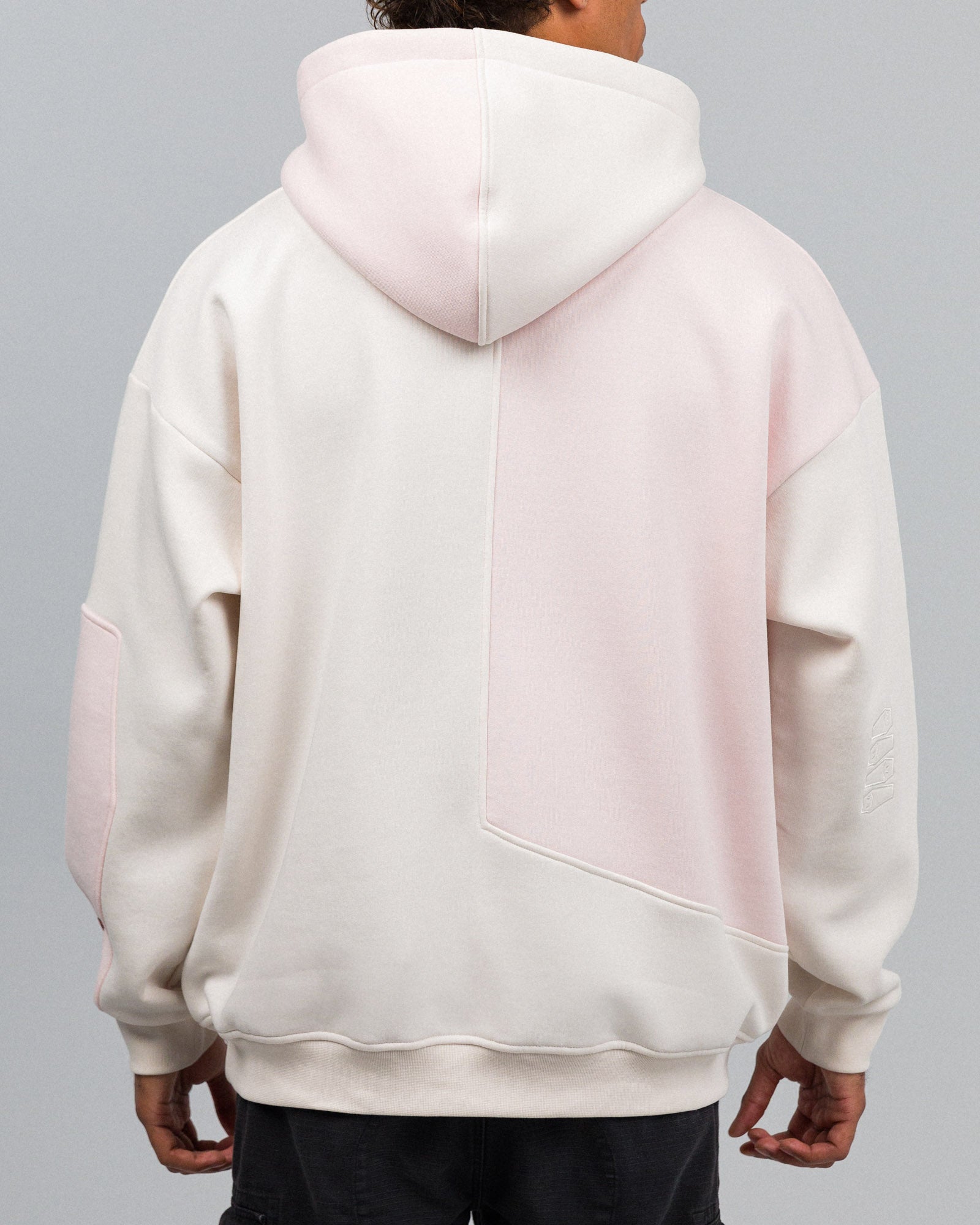 Rückseite des Hoodies mit großflächiger, asymmetrischer Teilung in Rosé und Cremeweiß. Die linke Seite ist cremefarben, die rechte zeigt einen großen rosa Einsatz. Die Kapuze ist ebenfalls zweifarbig.