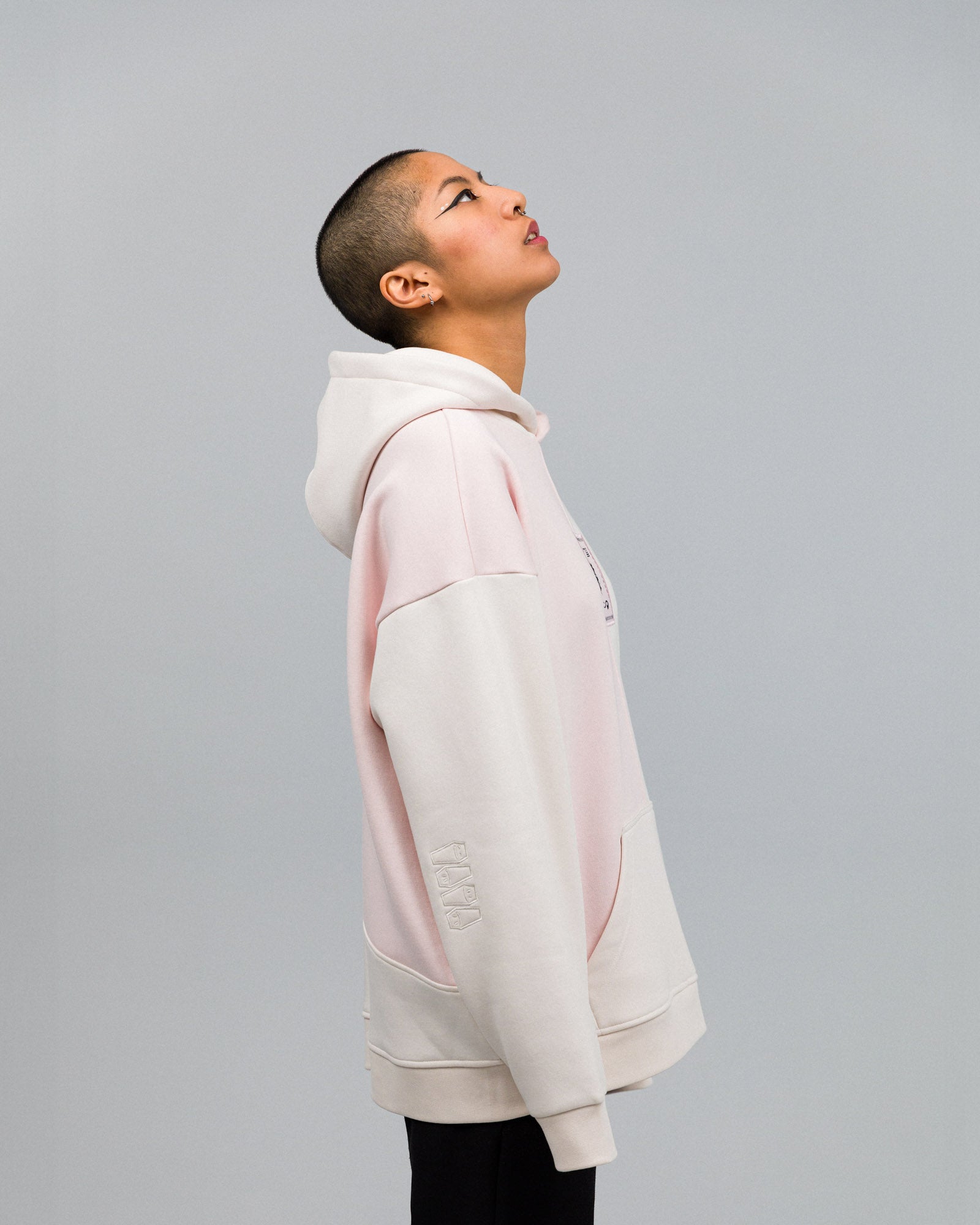 Seitenansicht eines Models im Hoodie. Die Teilung der Farbfelder in Rosé und Creme zieht sich über Ärmel, Kapuze und Rumpf. Die Stickerei mit den Sarg-Symbolen ist gut sichtbar am unteren Ärmel.