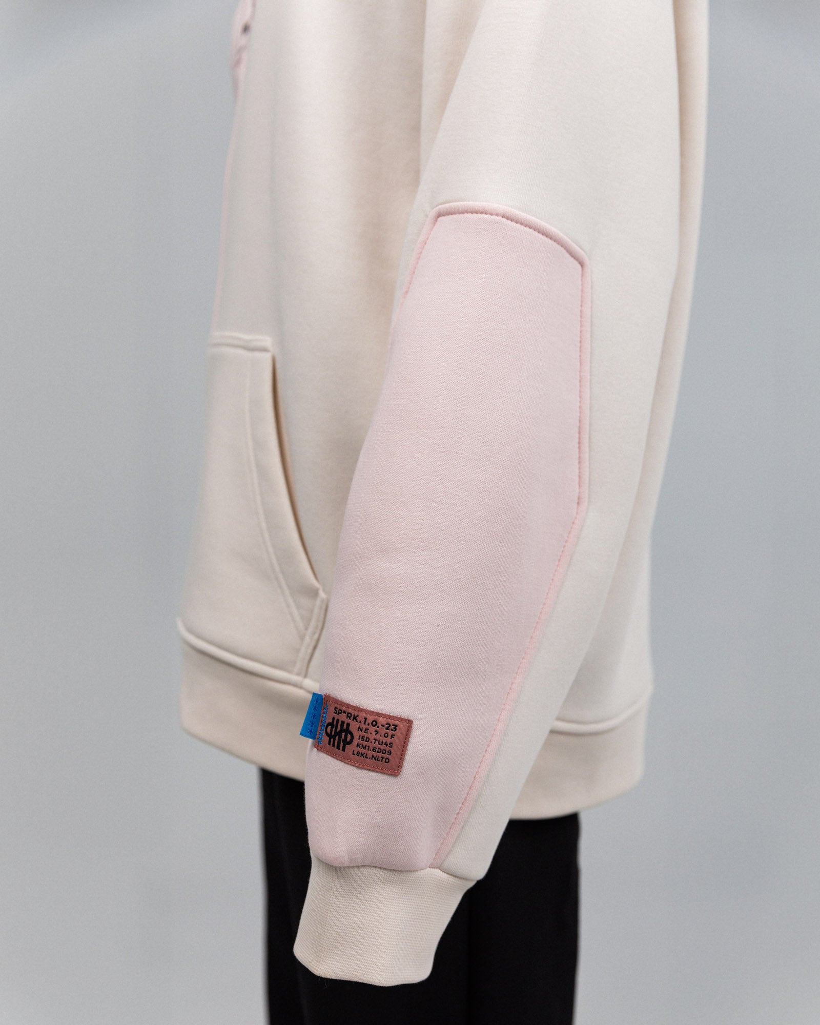 Rosafarbener Ellenbogenbereich mit aufgenähtem Stoffeinsatz. Am Bündchen ein Label mit blauer Lasche und braunem Patch: Text „SPRK 1.0.23“ sowie weitere kodierte Angaben.