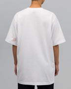 Rückansicht des weißen T-Shirts. Es ist schlicht und weit geschnitten. Auf dem linken Ärmel befindet sich ein kleiner roter Print mit Buchstaben- und Zahlenkombination.