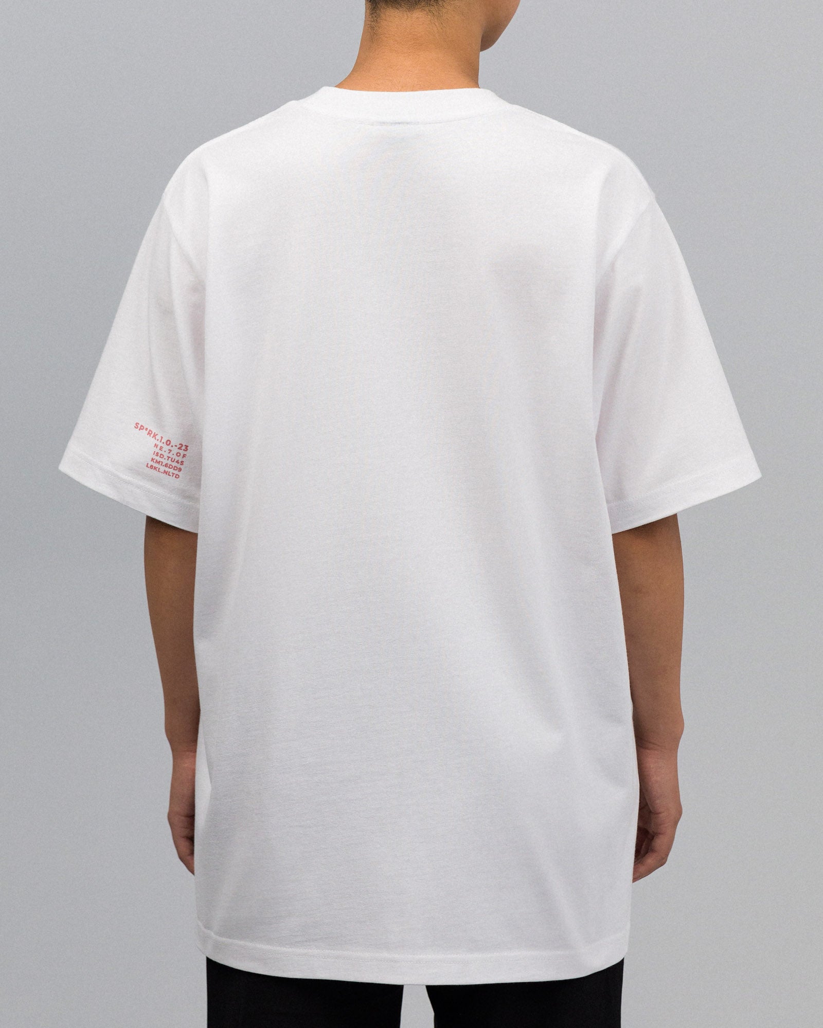 Rückansicht des weißen T-Shirts. Es ist schlicht und weit geschnitten. Auf dem linken Ärmel befindet sich ein kleiner roter Print mit Buchstaben- und Zahlenkombination.