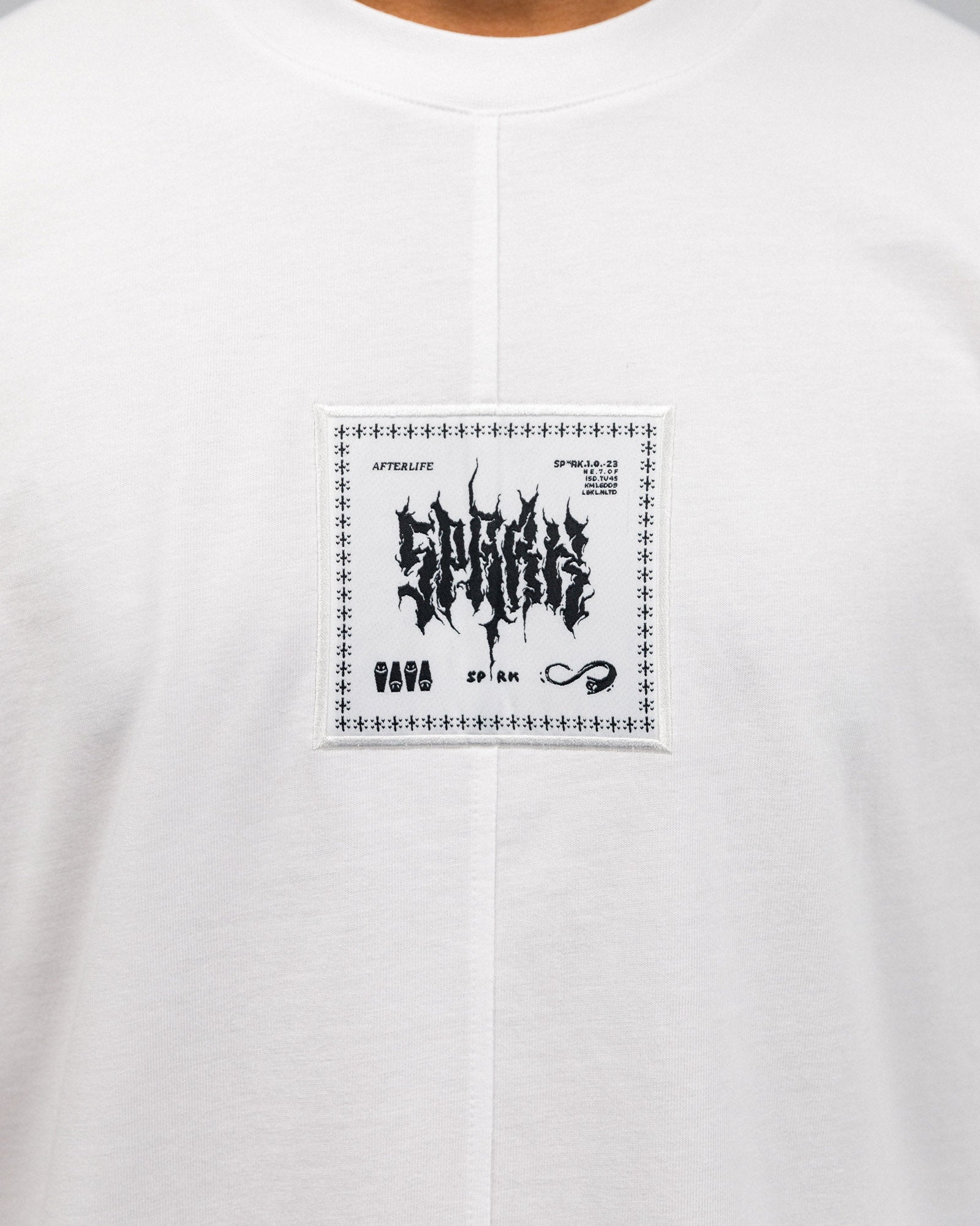 Detailansicht des schwarzen Frontpatches auf weißem Stoff. Im Zentrum steht das Wort „AFTERLIFE“ in verzerrter Typografie. Umrahmt von grafischen Zeichen, u. a. Symbolen wie Särgen und einer Unendlichkeitslinie.