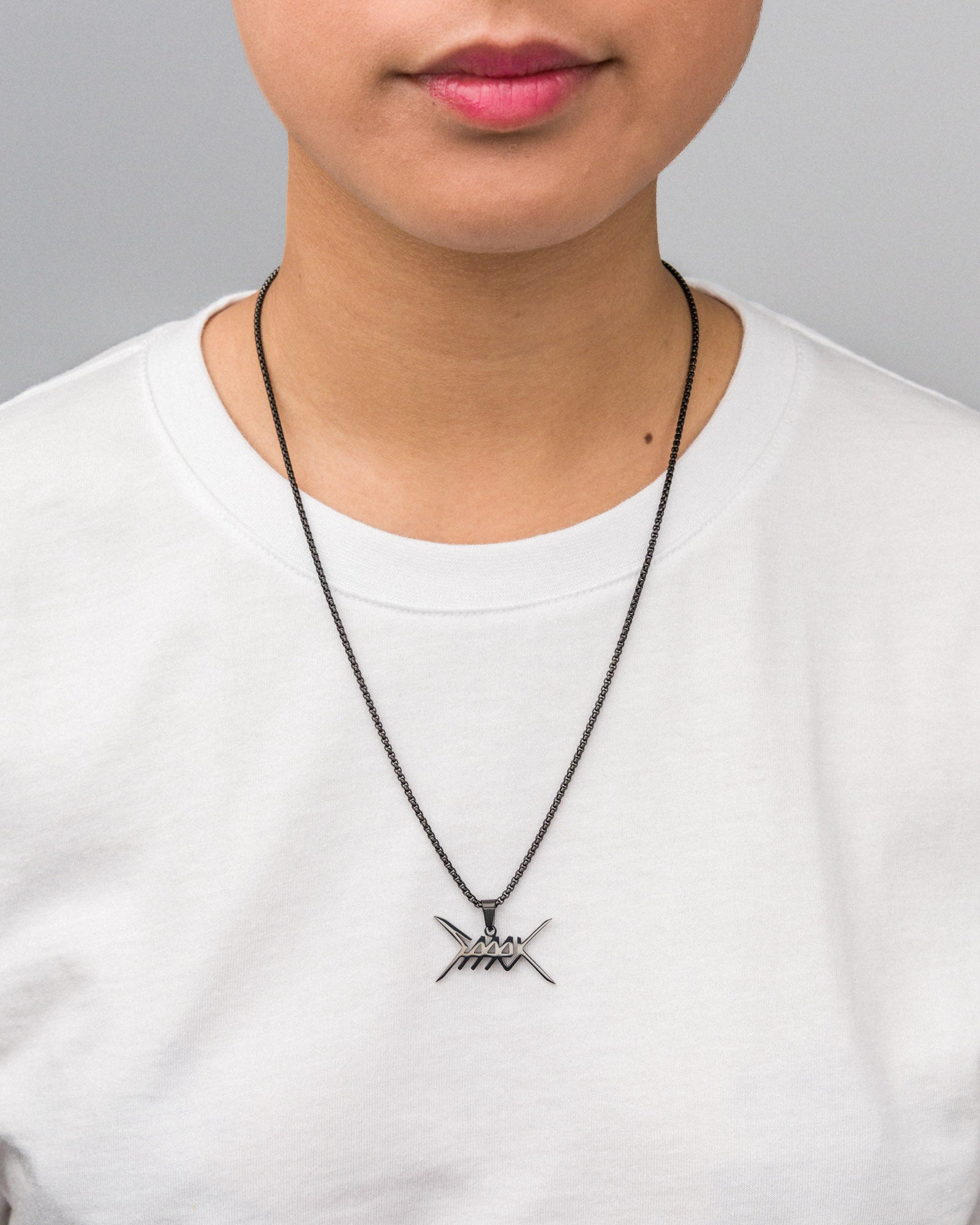 Person mit weißem T-Shirt trägt eine silberfarbene Halskette mit schwarzer Gliederkette; der mittig platzierte Anhänger zeigt ein metallisch glänzendes Logo in grafischer Stacheldraht-Anmutung.