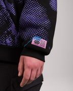 Linker Ärmel eines schwarzen Hoodies mit violettem Muster. Am Bündchen ist ein rosafarbener Label-Patch mit Zickzack-Naht und typografischen Elementen wie „HIP“ und technischen Angaben aufgenäht. Eine blaue Ziernaht verläuft quer über das Bündchen.