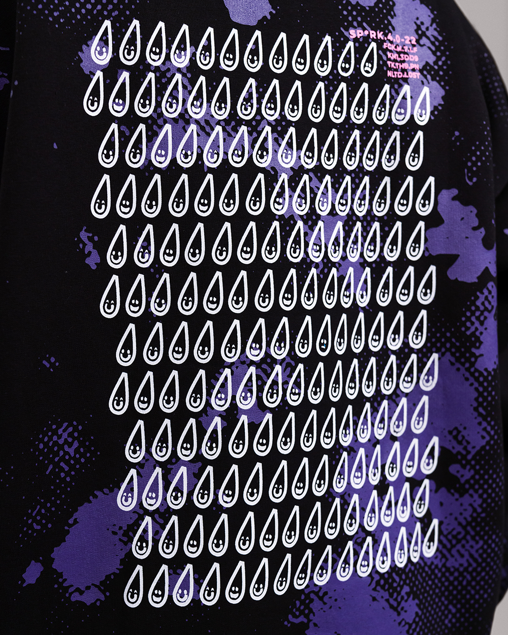 Großflächiger Print auf dem Rücken eines schwarzen Hoodies. Das Motiv besteht aus mehreren Reihen weißer Tropfen mit Gesichtern, auf violettem Punktmuster und Spritzflecken. Oben rechts befindet sich ein kleiner, orangefarbener Schriftzug mit kryptischen Zahlen- und Buchstabenkombinationen.