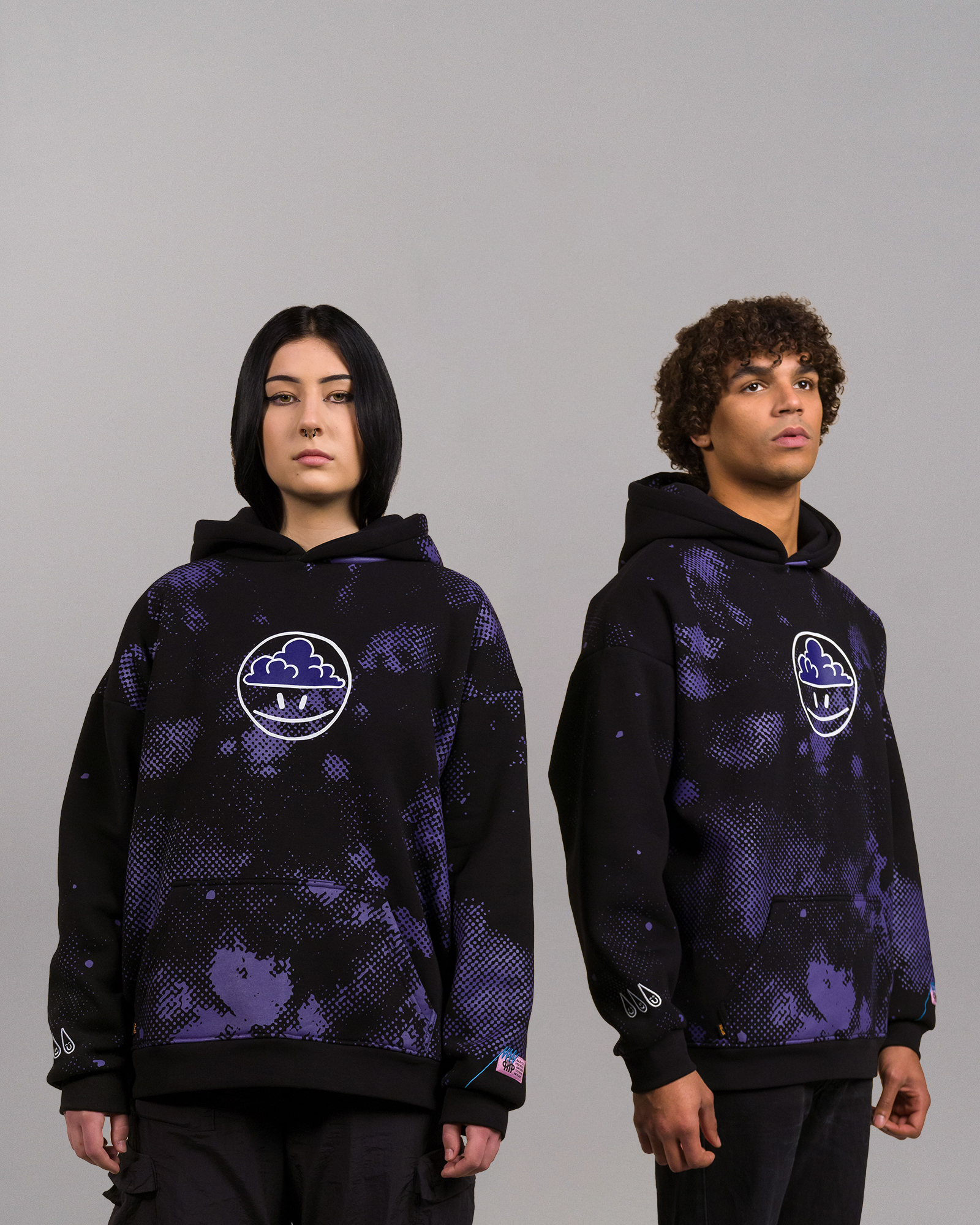 Zwei Personen tragen identische schwarze Hoodies mit lila Punktmuster und zentriertem Frontprint eines stilisierten weißen Gesichts mit Wolkenfrisur in einem Kreis. Beide Hoodies zeigen das Tropfensymbol am rechten Ärmel.