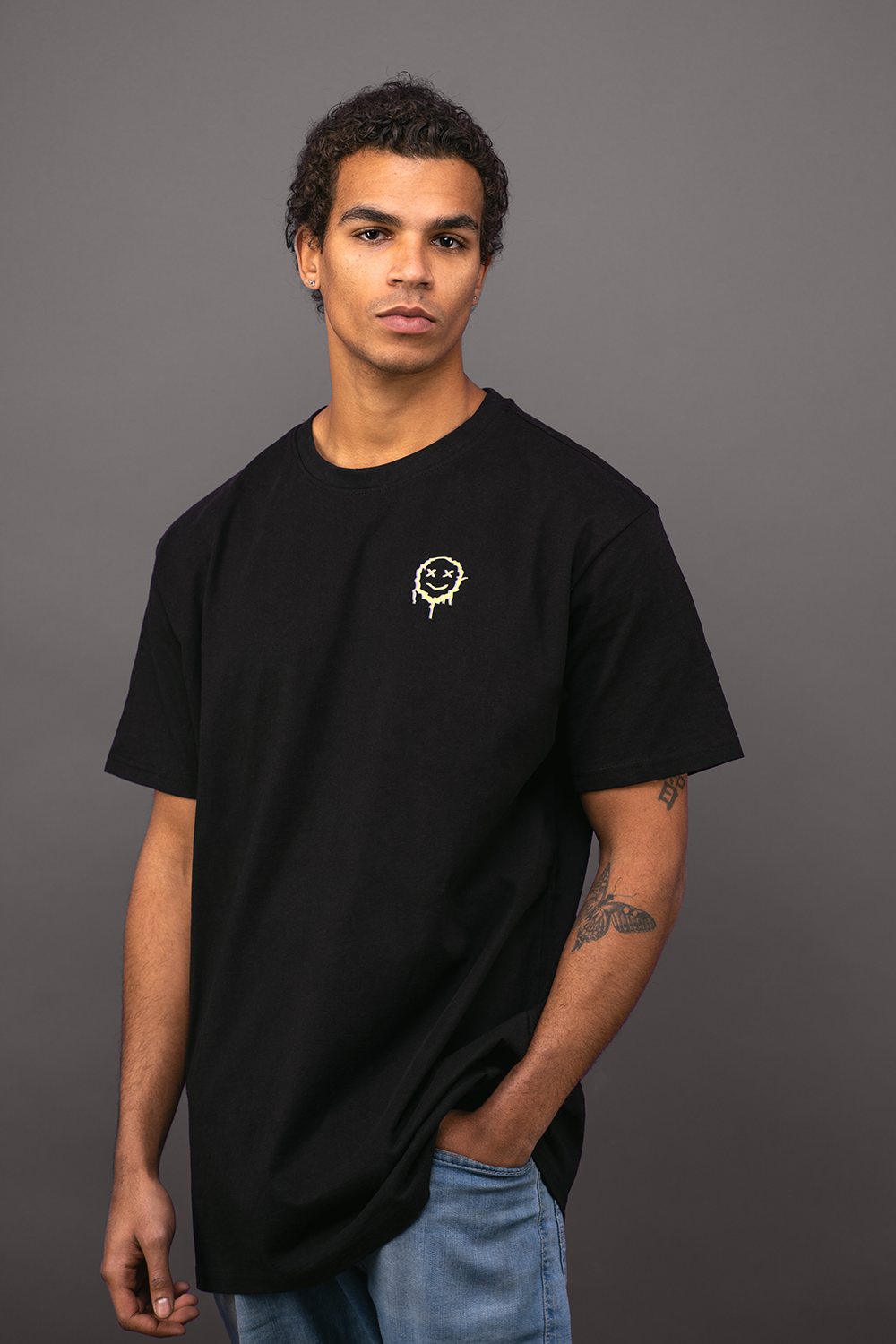 Schwarzes T-Shirt mit kleinem Frontprint auf Brusthöhe. Das Motiv zeigt ein gelbes, verlaufend gedrucktes Smiley-Gesicht mit gekreuzten Augen und tropfenartigem Verlauf nach unten. Die Person trägt das Shirt über hellblauer Jeans vor grauem Hintergrund.

