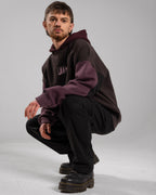 Hockende Pose einer Person im Studio. Der Fokus liegt auf dem locker getragenen Hoodie mit weinroten Ärmeln, rosa Frontlogo und dunkler Kängurutasche. Schwarze Hose und Schuhe runden das Outfit ab.

