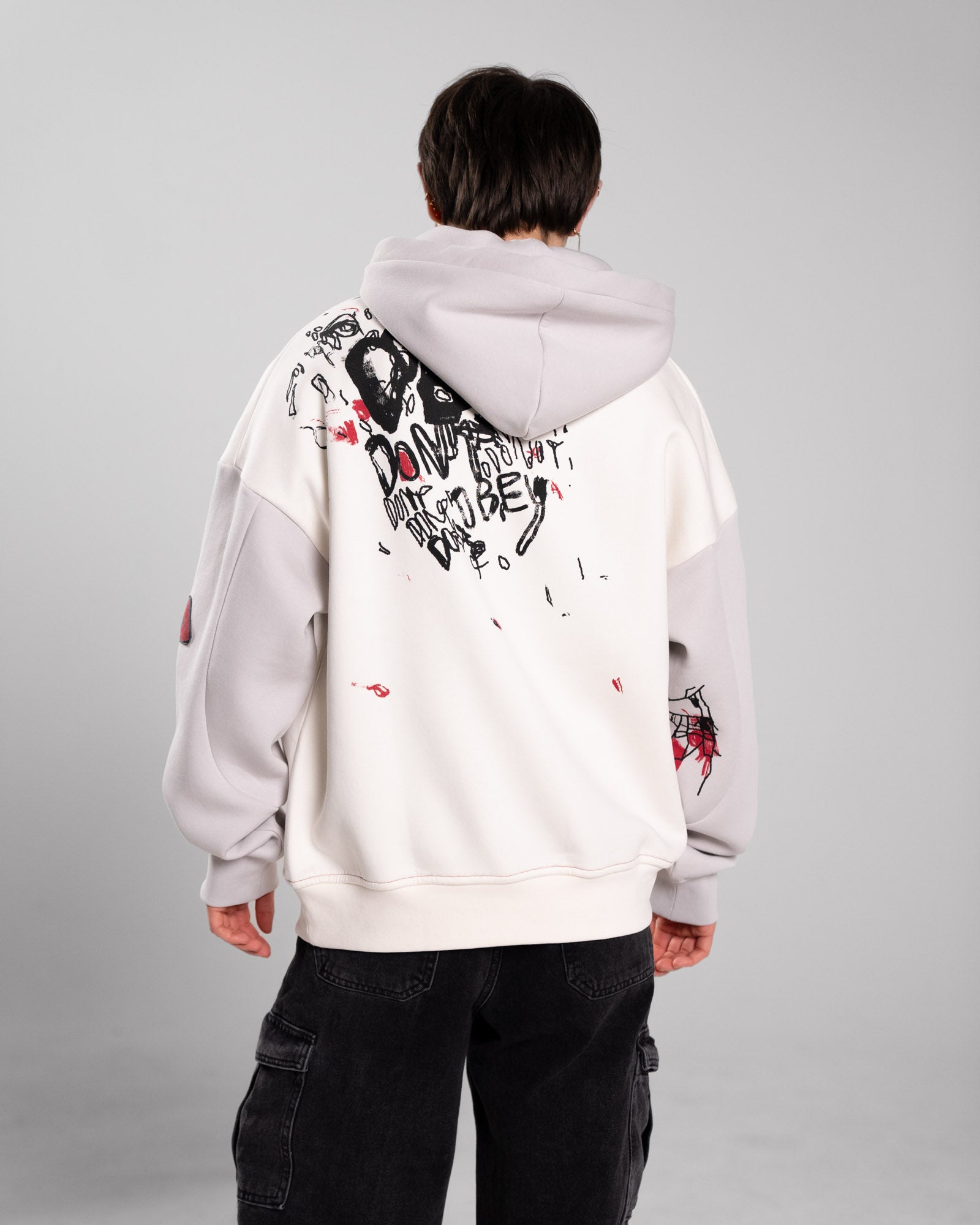 Rückansicht des Hoodies mit auffälligem schwarz-rotem Rückendruck. Der Print zeigt Kritzelgrafiken, Gesichter und den handschriftlich wirkenden Schriftzug „DON’T OBEY“ mehrfach wiederholt. Auf dem Ärmel sind rote und schwarze Linienzeichnungen sichtbar.