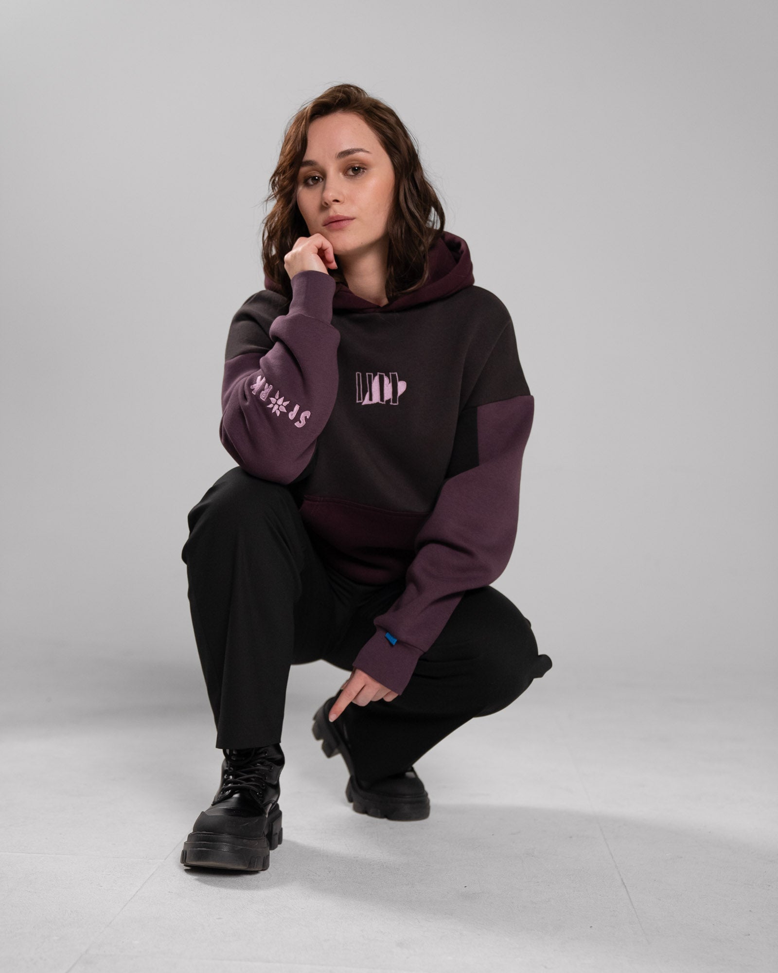 Hockende Pose einer Person im Studio. Der Fokus liegt auf dem locker getragenen Hoodie mit weinroten Ärmeln, rosa Frontlogo und dunkler Kängurutasche. Schwarze Hose und Schuhe runden das Outfit ab.

