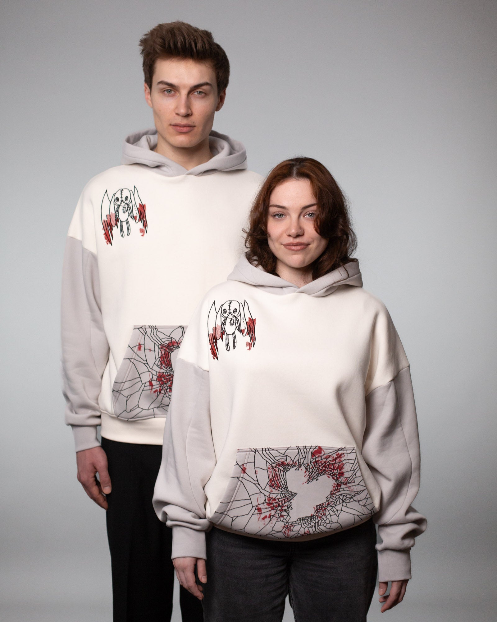 Zwei Personen tragen denselben cremefarbenen Hoodie mit grauen Ärmeln und Kapuze. Die Vorderseite des Hoodies zeigt links auf der Brust eine gestickte Illustration eines geflügelten Stoffwesens mit roten Akzenten. Die Kängurutasche ist mit einem schwarzen Spinnennetzmuster bedruckt, das eine zentrale herzförmige Aussparung freilässt, ergänzt durch rote Spritzmuster. Beide Models stehen nebeneinander in neutraler Studioumgebung.