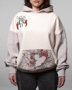 Vorderansicht des cremefarbenen Hoodies mit grauen Ärmeln und Kapuze. Links auf der Brust ist eine schwarze Illustration einer geflügelten Stofffigur mit roten Akzenten aufgestickt. Die Kängurutasche zeigt ein zersplittertes Spinnennetz-Muster mit herzförmiger Aussparung und roten Spritzern.