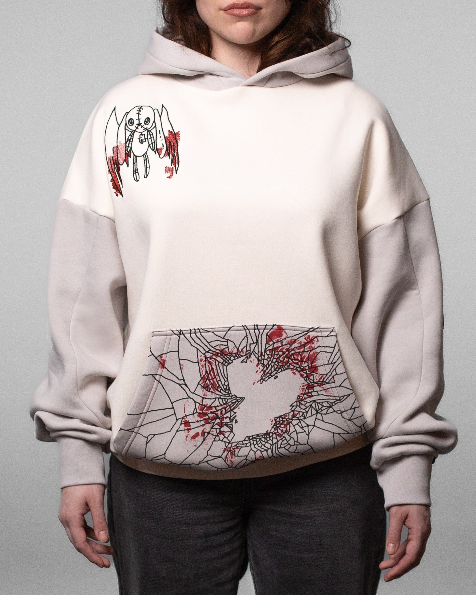 Vorderansicht des cremefarbenen Hoodies mit grauen Ärmeln und Kapuze. Links auf der Brust ist eine schwarze Illustration einer geflügelten Stofffigur mit roten Akzenten aufgestickt. Die Kängurutasche zeigt ein zersplittertes Spinnennetz-Muster mit herzförmiger Aussparung und roten Spritzern.