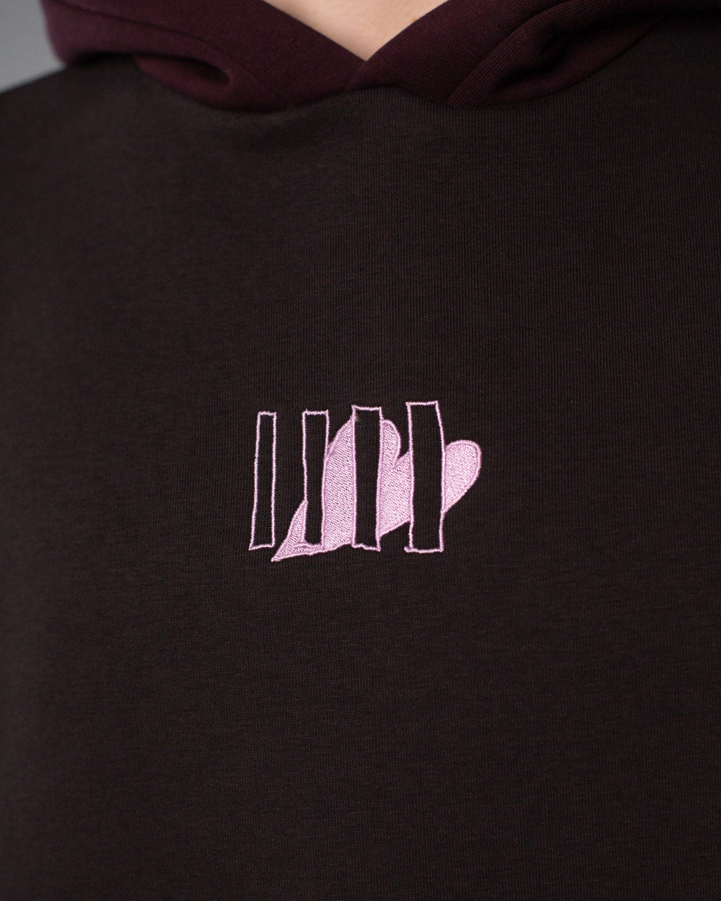Detailaufnahme des Brustlogos: fünf senkrechte Linien mit integrierter Herzform, aufgestickt in hellem Rosa auf dunkelbraunem Stoff.