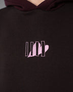 Detailaufnahme des Brustlogos: fünf senkrechte Linien mit integrierter Herzform, aufgestickt in hellem Rosa auf dunkelbraunem Stoff.