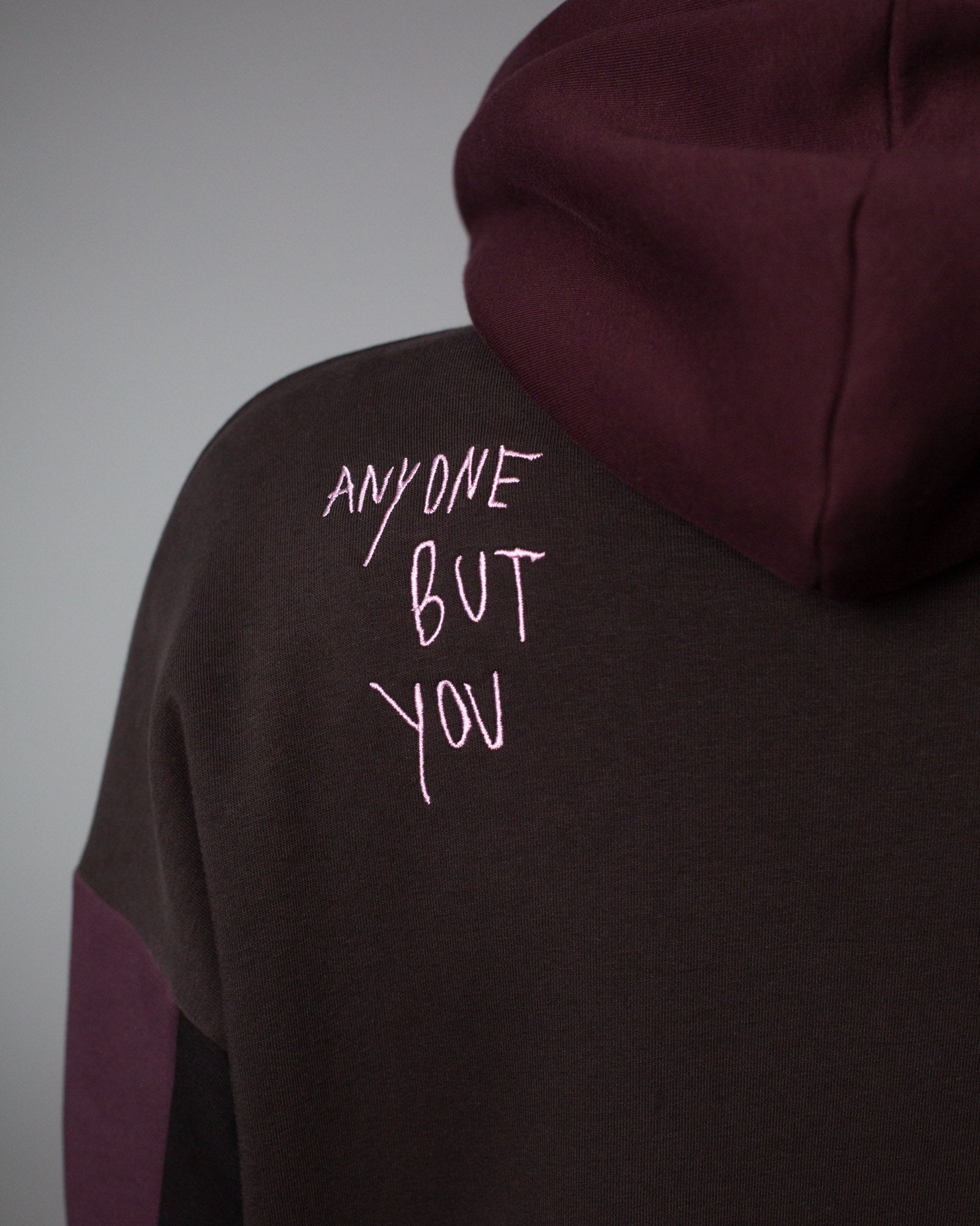 Nahansicht der Rückseite des Hoodies mit sichtbarer Kapuze in Weinrot und dem hellrosa gestickten Schriftzug „ANYONE BUT YOU“ im Nackenbereich.