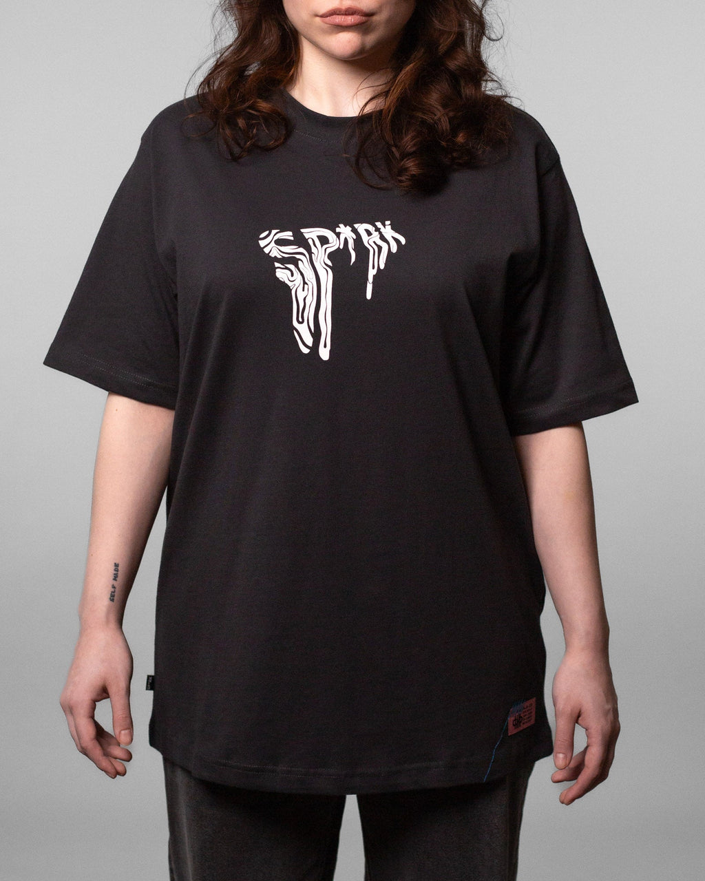 Frontalaufnahme einer Frau im schwarzen T-Shirt mit weißem „SPRK“-Brustprint. Der Schnitt fällt locker und reicht über die Hüfte.
