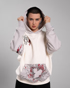 Frontalansicht eines Models mit hochgezogener Kapuze. Sichtbar sind die Illustration der geflügelten Stofffigur auf der Brust, die grafische Kängurutasche sowie rote und schwarze Druckdetails am Ärmel.