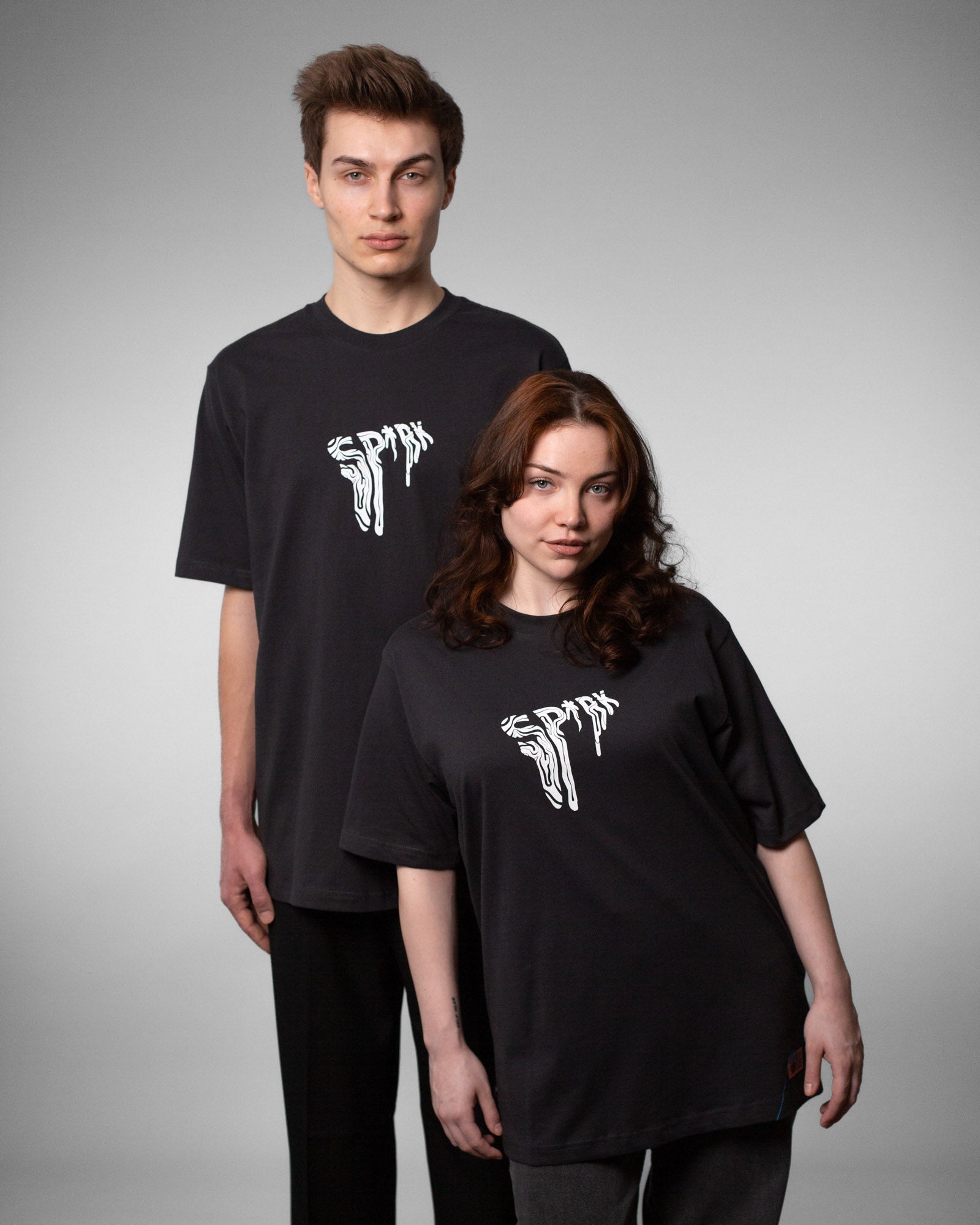 Zwei Personen in schwarzen T-Shirts mit identischem weißen „SPRK“-Brustlogo. Die linke Person trägt das Shirt oversized, die rechte in regulärer Passform. Beide stehen in neutraler Studio-Umgebung.