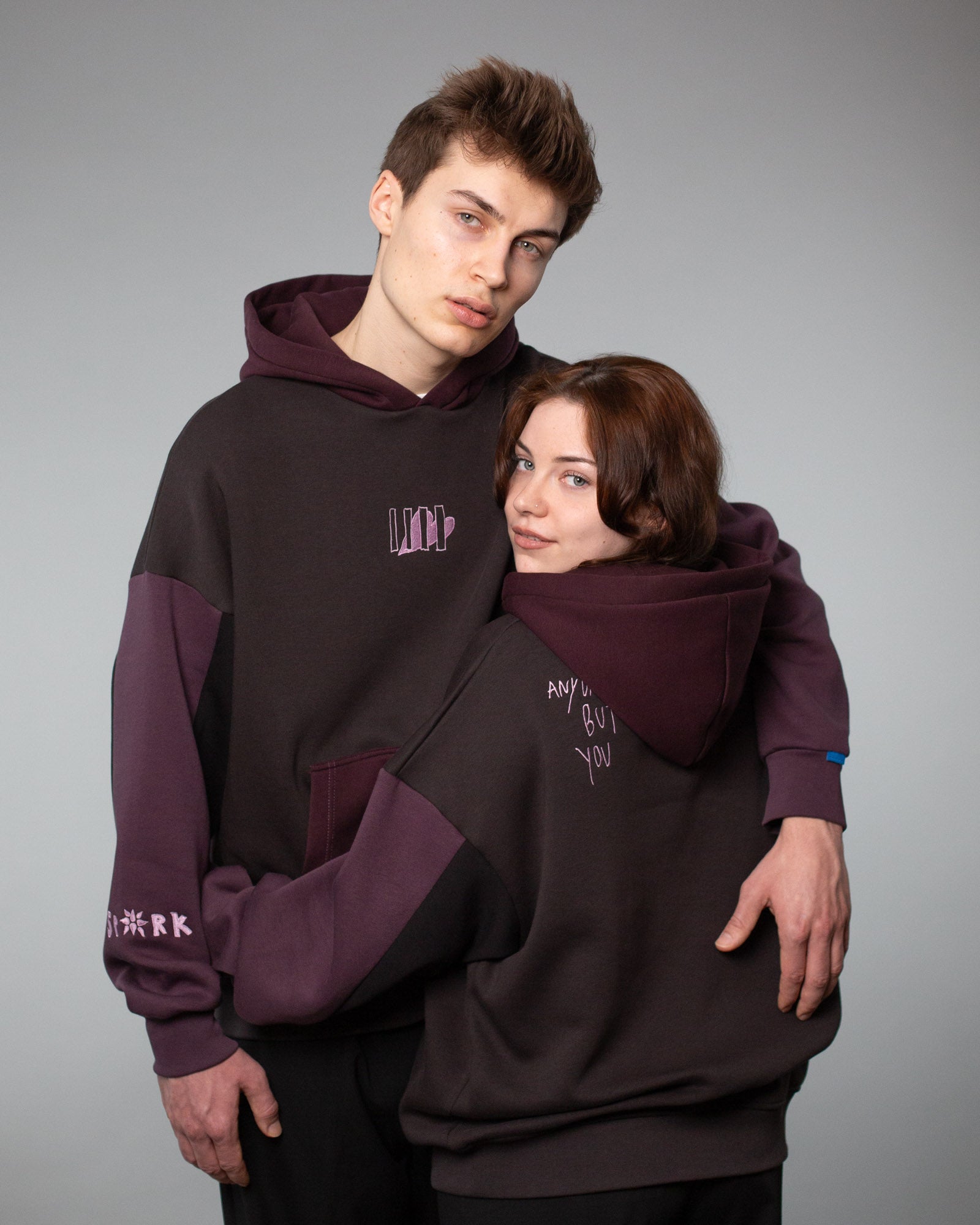 Zwei Personen tragen denselben zweifarbigen Hoodie in dunklem Braun und Weinrot. Die Person im Vordergrund zeigt die Rückenpartie mit der Stickerei „ANYONE BUT YOU“, während die andere Person frontal steht und die Ärmelstickerei „SPARK“ sichtbar ist.