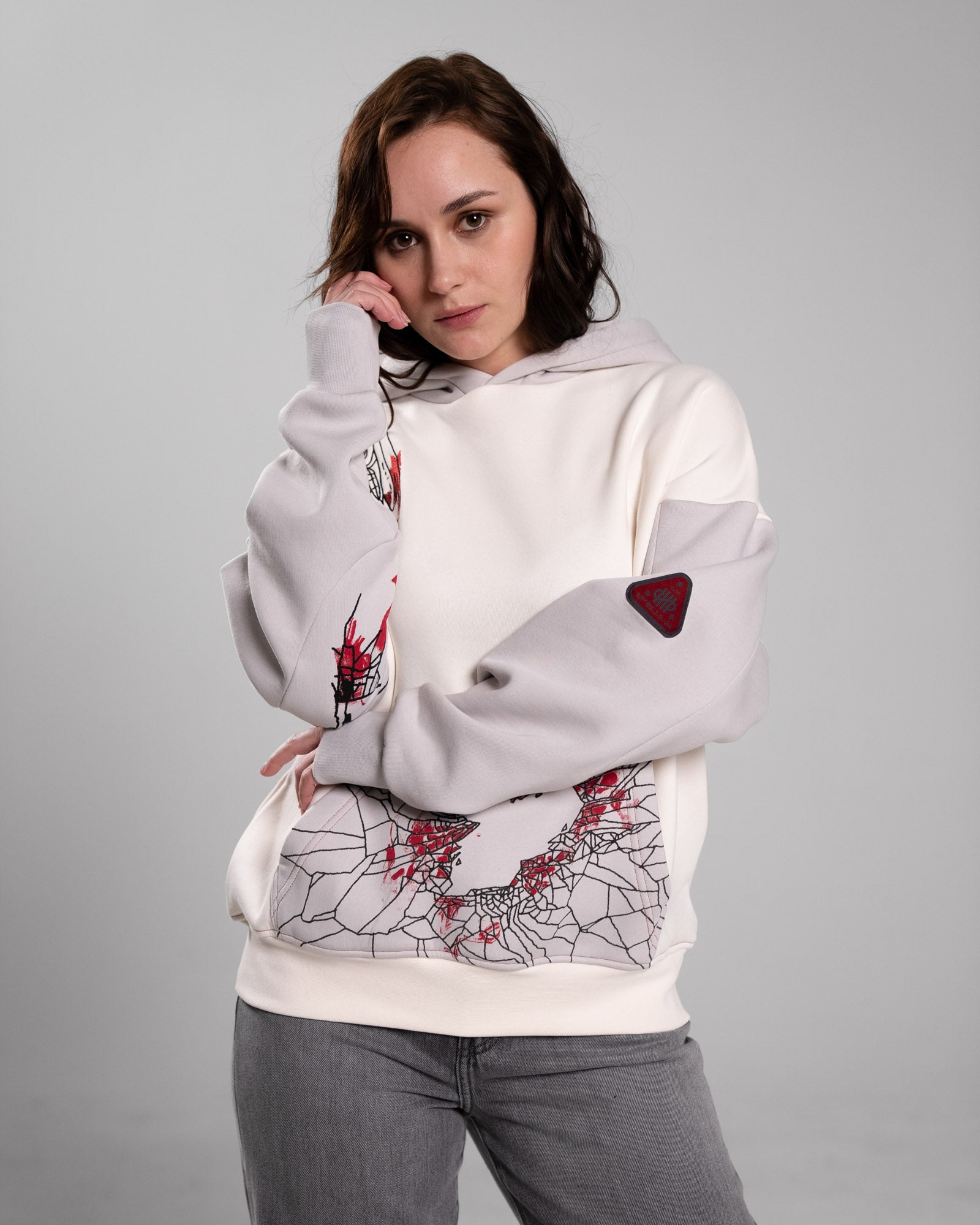 Frontalaufnahme einer Person mit langem Haar im Hoodie. Der Fokus liegt auf dem roten Ärmelpatch, der Brustillustration und der Kängurutasche mit grafischem Herzmotiv.